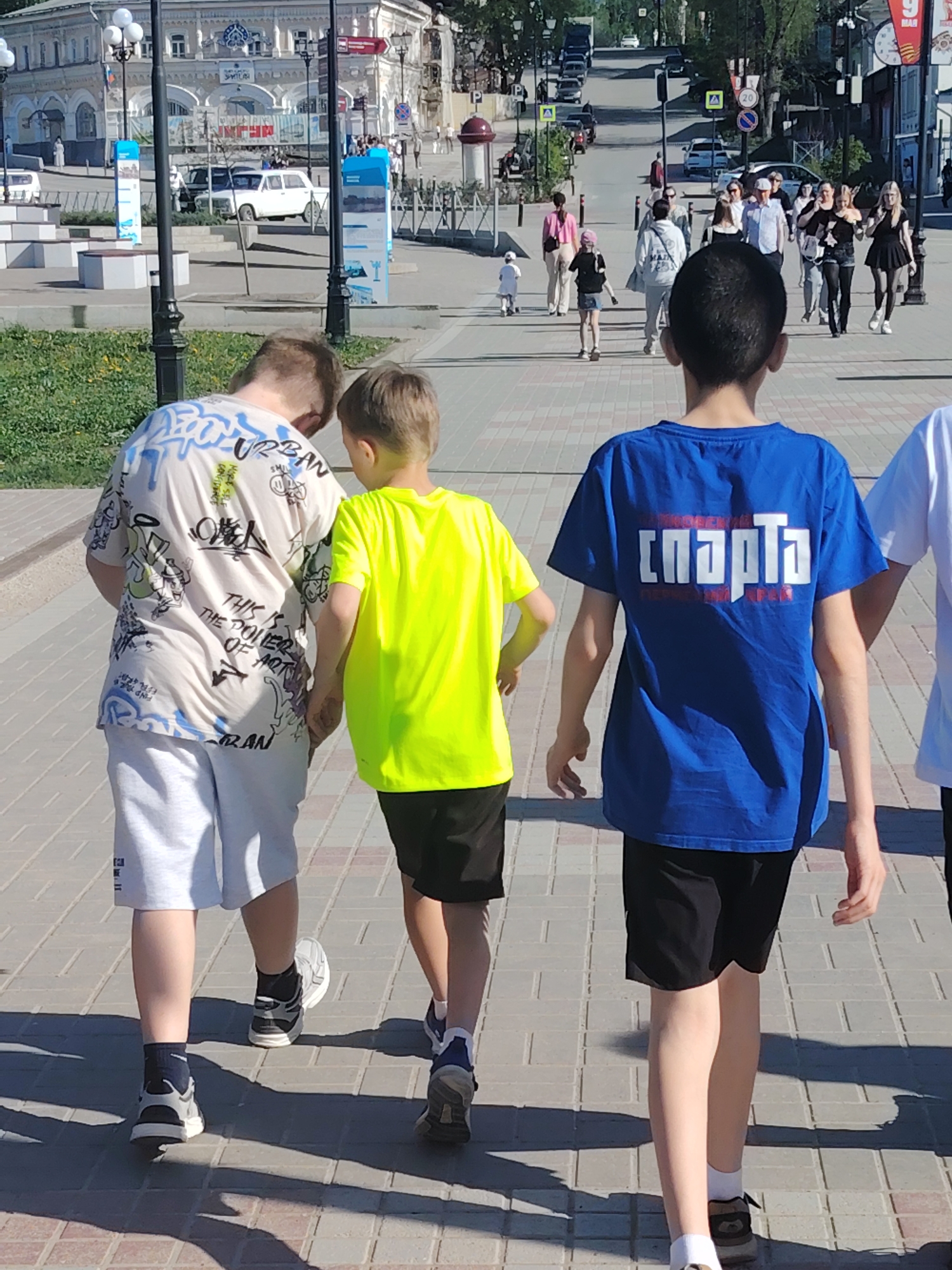 On a sunny day in Kungur, Russia, three boys in casual attire walk along a paved brick pathway. The first boy wears a white, graphic t-shirt with light-colored shorts and gray sneakers with black socks. The second boy is dressed in a bright neon-yellow t-shirt and black shorts with gray sneakers. The third boy wears a blue t-shirt with the Russian word "спарта" printed on the back, paired with black shorts and white socks and sneakers. The street is populated with pedestrians in the background. The first buildings appear to be two-story structures. Vehicles are parked on the street. A building with a clock tower can be seen. The atmosphere suggests a casual outing in an urban setting.