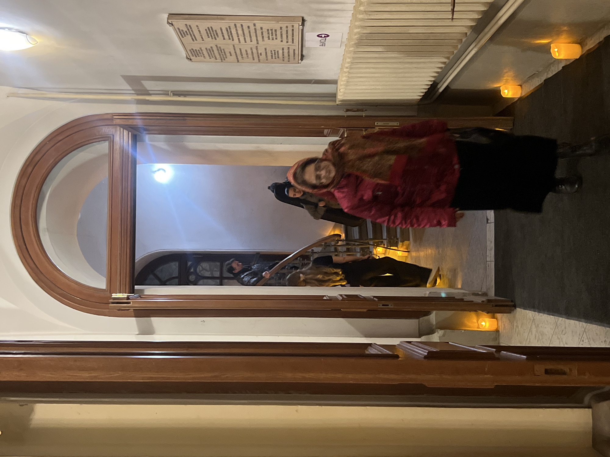 The scene is an interior shot of a building with architectural details suggestive of Saint Petersburg, Russia. The photograph is taken inside a doorway with a large, arched wooden frame. The lighting is mixed, with an ambient glow and what appears to be additional lighting elements positioned within the space, some on the floor.
A woman in a red jacket and a headscarf is walking towards the camera, slightly off-center. Behind her, other people are descending a short flight of steps. The people in the background are not clearly discernible.
To the left, another person is visible inside the doorway.
Various signs and plaques can be seen on the walls, as well as a radiator.
