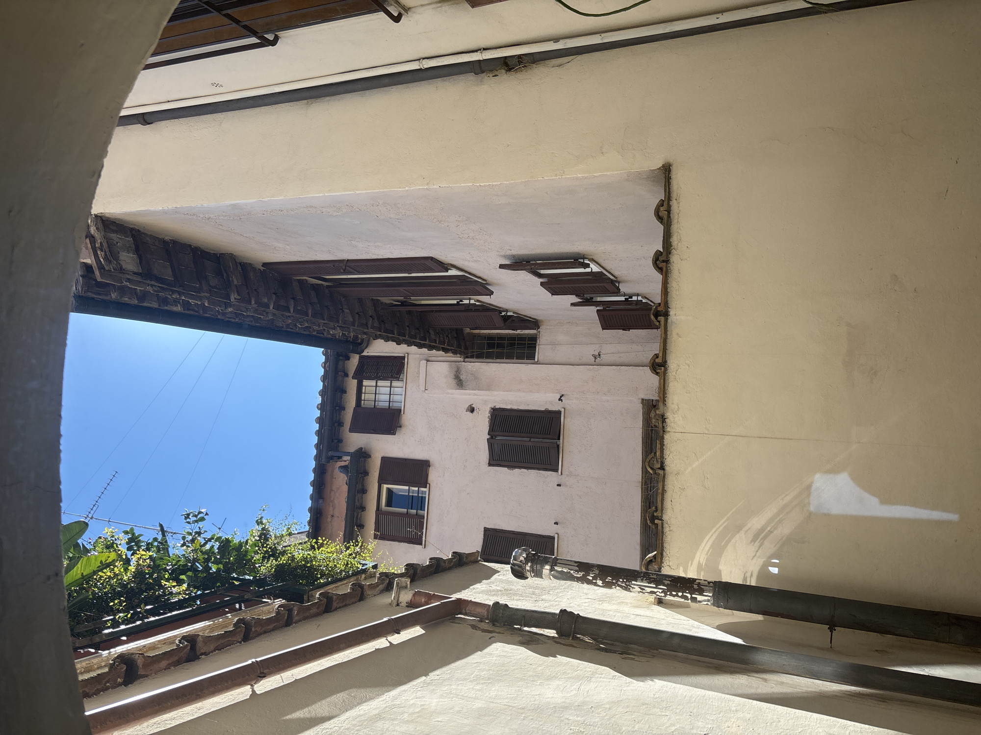The image shows an upward view from a courtyard in Rome, Italy. The architecture features light-colored walls and terracotta roof tiles. Brown shutters are visible on the windows of a building opposite the vantage point. Green foliage grows atop a lower roof, with a clear blue sky visible above. Metal pipes run along the building surfaces.
