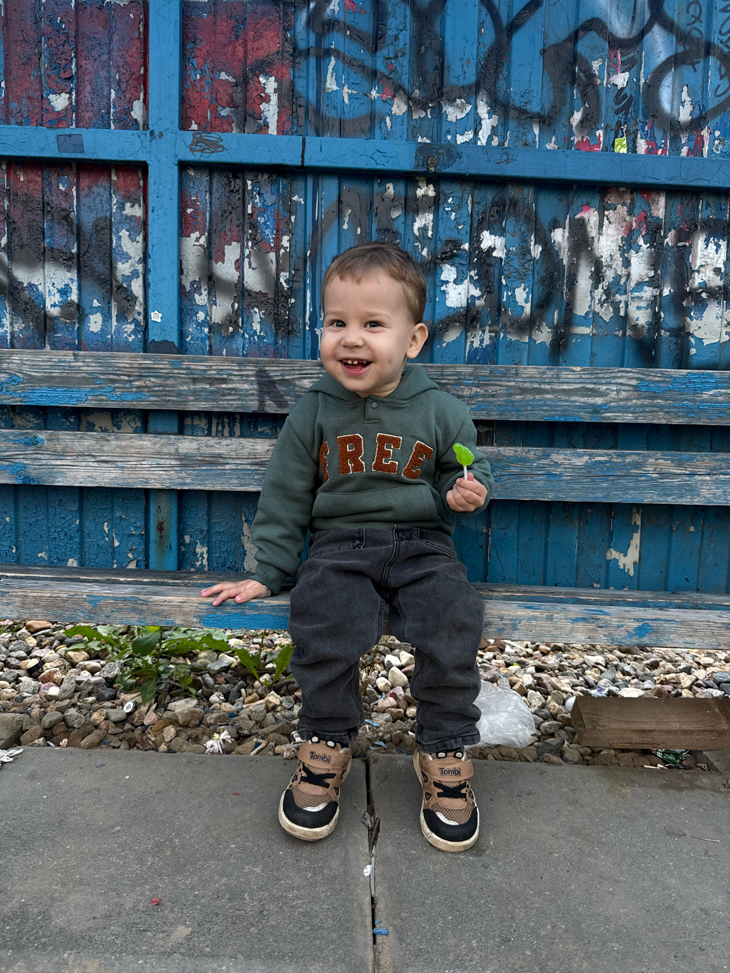 A young boy is sitting on a weathered wooden bench in front of a blue, graffiti-covered wall in Yaroslavl, Russia. He is wearing a green hoodie with the word "FREE" in orange letters, dark gray jeans, and brown and black sneakers. The boy is smiling and holding a green lollipop in his right hand. The bench is made of rough wooden planks, and the ground in front of it is covered in gravel and some debris, including a plastic bag and a piece of wood. The wall behind the bench is blue with various peeling layers of paint, revealing red and white underneath, and is extensively covered in black and blue graffiti tags and scribbles. The overall scene appears to be in an outdoor urban setting.