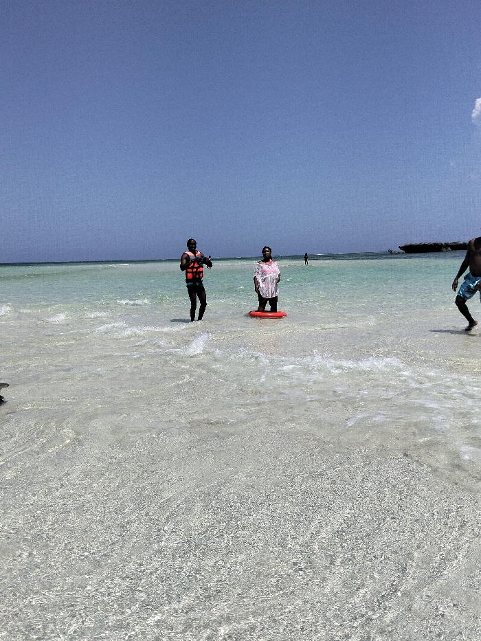 The image depicts a bright, clear day on a shallow coastal area in Wasini, Kenya. Three individuals are prominently featured in the turquoise-green ocean water with a visible light-colored, sandy seabed.
On the left, an adult male with dark skin is standing in the water, wearing dark-colored bottoms and an orange life vest. He is holding a dark object, possibly a camera or phone, at chest level, and is angled slightly towards the right.
In the center, an adult female with dark skin is standing in the water. She is wearing a light-colored top over a pink garment and has an orange inflatable ring around her waist. She is facing forward.
On the far right, partially visible, another adult male with dark skin is stepping through the water, wearing light blue and white patterned swim shorts.
In the background, the clear blue sky meets the horizon where the ocean extends. Distant figures are visible further out in the water, and a dark, rocky outcrop or small island is present on the right horizon. Small waves are visible around the subjects.