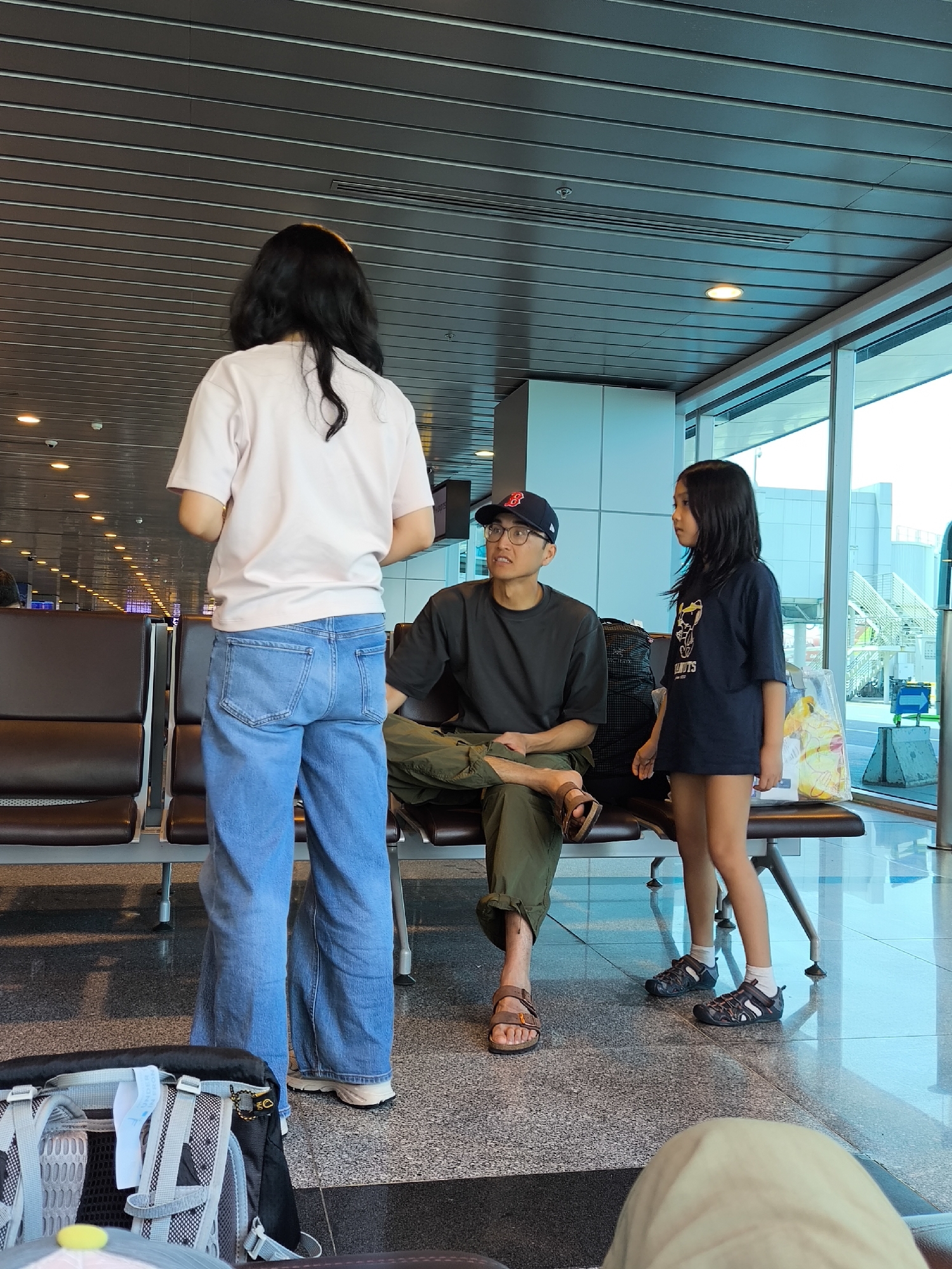The scene is located within the passenger terminal of Đà Nẵng International Airport, Vietnam. Three individuals are present: two adults and one child.
An adult female stands on the left, viewed from behind. She has long dark hair and wears a light pink short-sleeved t-shirt and light blue wide-leg jeans, with white sneakers partially visible. She faces towards the right.
An adult male is seated on a dark brown multi-seat airport waiting bench in the center. He wears a dark baseball cap with a red 'B' logo, eyeglasses, a dark short-sleeved t-shirt, olive green cargo pants rolled above the ankles, and brown leather sandals. His legs are crossed at the ankles. He looks upward, towards the standing female. A black and grey Arc'teryx backpack rests on the dark speckled terrazzo floor next to his left foot.
To the right of the seated male, a pre-adolescent female child stands. She has long dark hair and wears a dark blue short-sleeved t-shirt with a white graphic (resembling Peanuts characters) and dark closed-toe sandals with white socks. She faces towards the large, floor-to-ceiling windows on the right, which overlook an airport tarmac. Outside, a light-colored building with white external stairs and railings, and a blue luggage cart, are visible under a bright sky. A white plastic shopping bag with yellow graphics is placed on the waiting chair beside the child.
The ceiling consists of parallel grey metallic panels with recessed circular lighting fixtures. The overall lighting is bright and consistent throughout the terminal area. A portion of another backpack is visible in the bottom-left foreground.