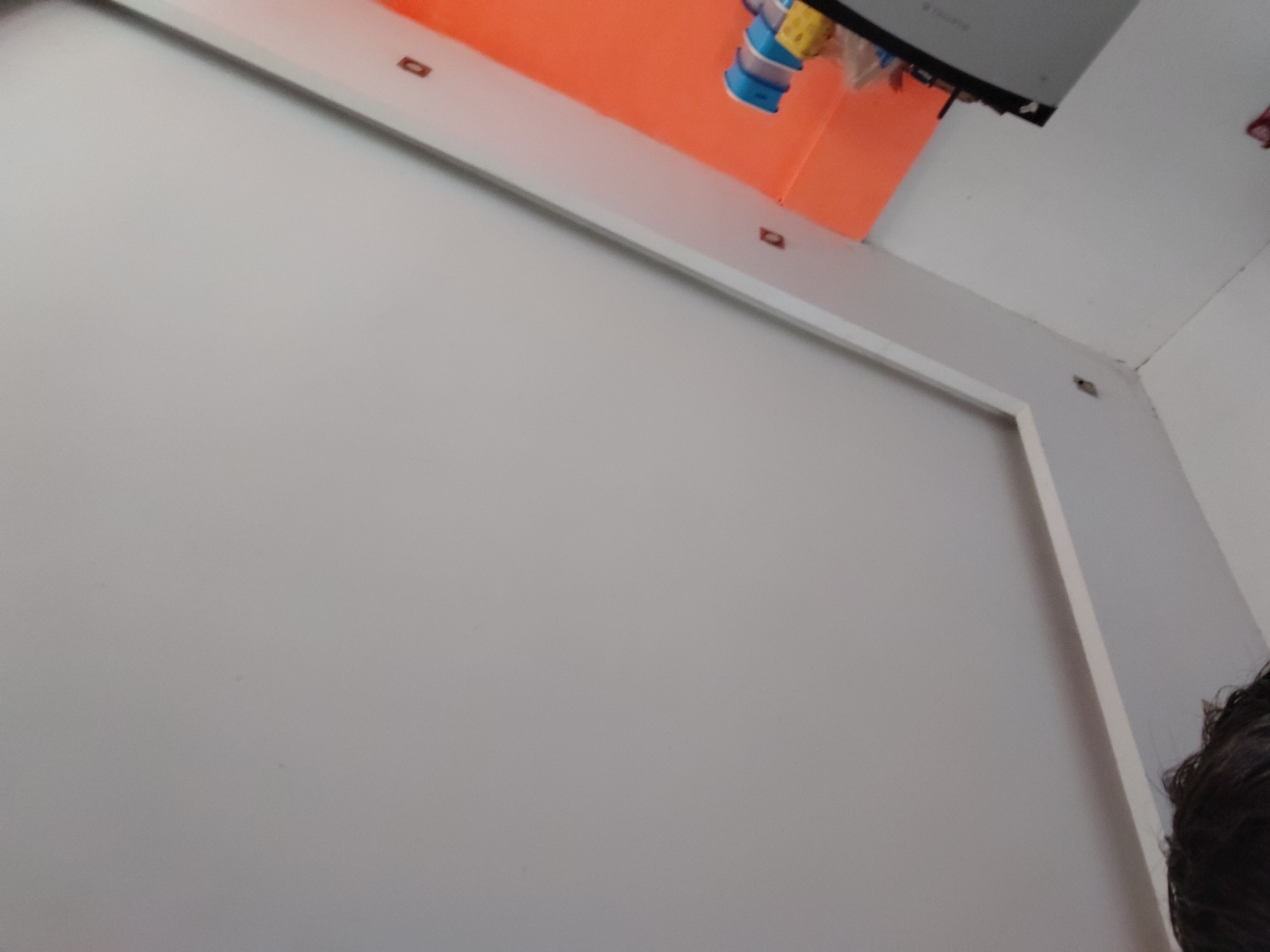 The image displays an indoor scene, likely a room in Santa Ana, Venezuela, captured from a low, upward angle. A large, plain white surface dominates the left and center, appearing to be a wall or screen. Above this white surface, a narrow white strip runs horizontally, featuring several small, dark, rectangular inserts or fasteners. Above this strip, an expanse of bright orange material or paint is visible, suggesting a vibrant ceiling or upper wall section.
In the top right corner, a portion of a gray and black object, possibly a laptop or screen, is partially visible, with faint, illegible text on its surface. Hanging down from above it is a blue and yellow object, possibly a toy or decoration. The bottom right corner shows a small section of dark, curly hair, indicating the presence of a person taking the photo or standing nearby. The lighting appears to be diffused, and no specific activity is clearly depicted.