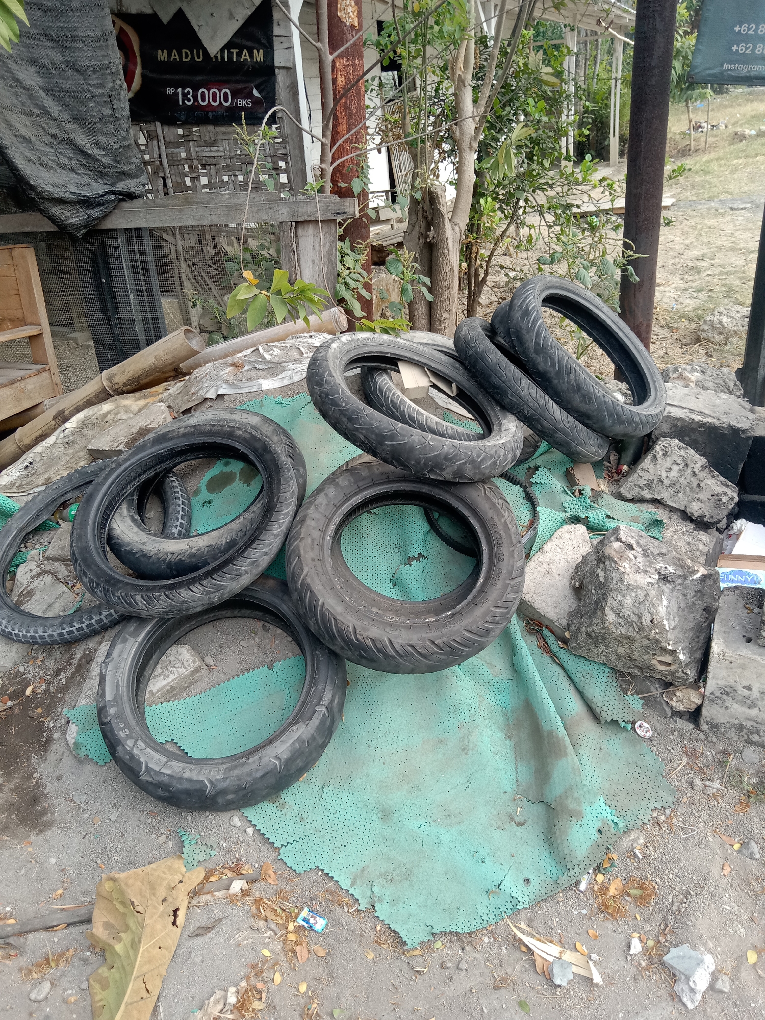 In Siwalan, Indonesia, a pile of used tires is stacked on a torn green mat. The tires, various sizes and brands, are scattered among rocks and debris on a dirt and gravel surface. In the background, a wooden structure with a mesh screen is visible, along with a sign displaying "MADU HITAM" (Black Honey) and a price of "RP 13.000/BKS". Trees and foliage surround the area. A metal pole stands to the right of the image. The scene suggests an outdoor storage or disposal area.