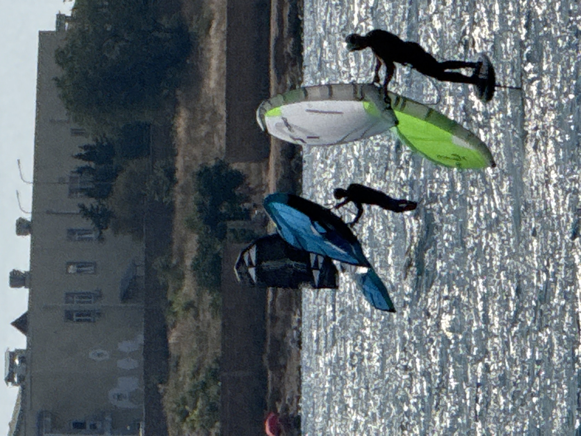 The image captures a bright, sunny day with choppy, sun-dappled water. In the foreground, three individuals are engaged in watersports. The person on the right is foilboarding with a kite. They are facing left, holding onto a kite bar and wearing dark clothing. Their foilboard is visible beneath them. To the left of this person, another individual is also foilboarding, facing forward and wearing dark clothing. They are holding a kite that is mostly out of frame but appears to be blue. In the middle distance, a dark kite is visible, with a person silhouette behind it, also seemingly engaged in a watersport.
In the background, a long, dark retaining wall runs horizontally across the image. Behind the wall, dry, scrubby vegetation covers a hillside. Further in the background, a large, light-colored building with a flat roof and multiple windows is situated on a slightly elevated plateau. The building appears to be multi-story and has some visible architectural details like chimneys and what might be vents or rooftop structures. Trees are sparsely scattered around the building and on the hillside. The overall scene suggests a coastal or bay area, potentially within Sevastopol, Ukraine, known for its naval history and watersports.