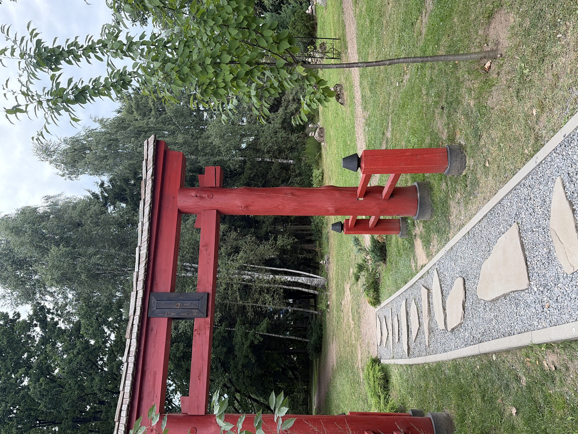 A red wooden torii gate stands to the left of the frame, partially obscuring a path of stepping stones. The path, made of large, flat stones embedded in gravel, leads away from the gate into a park-like setting with a lawn and trees. A small, slender tree with green leaves grows on the right side of the path. In the background, a dense forest of various trees, including some with white bark, creates a natural backdrop. The sky is overcast.
