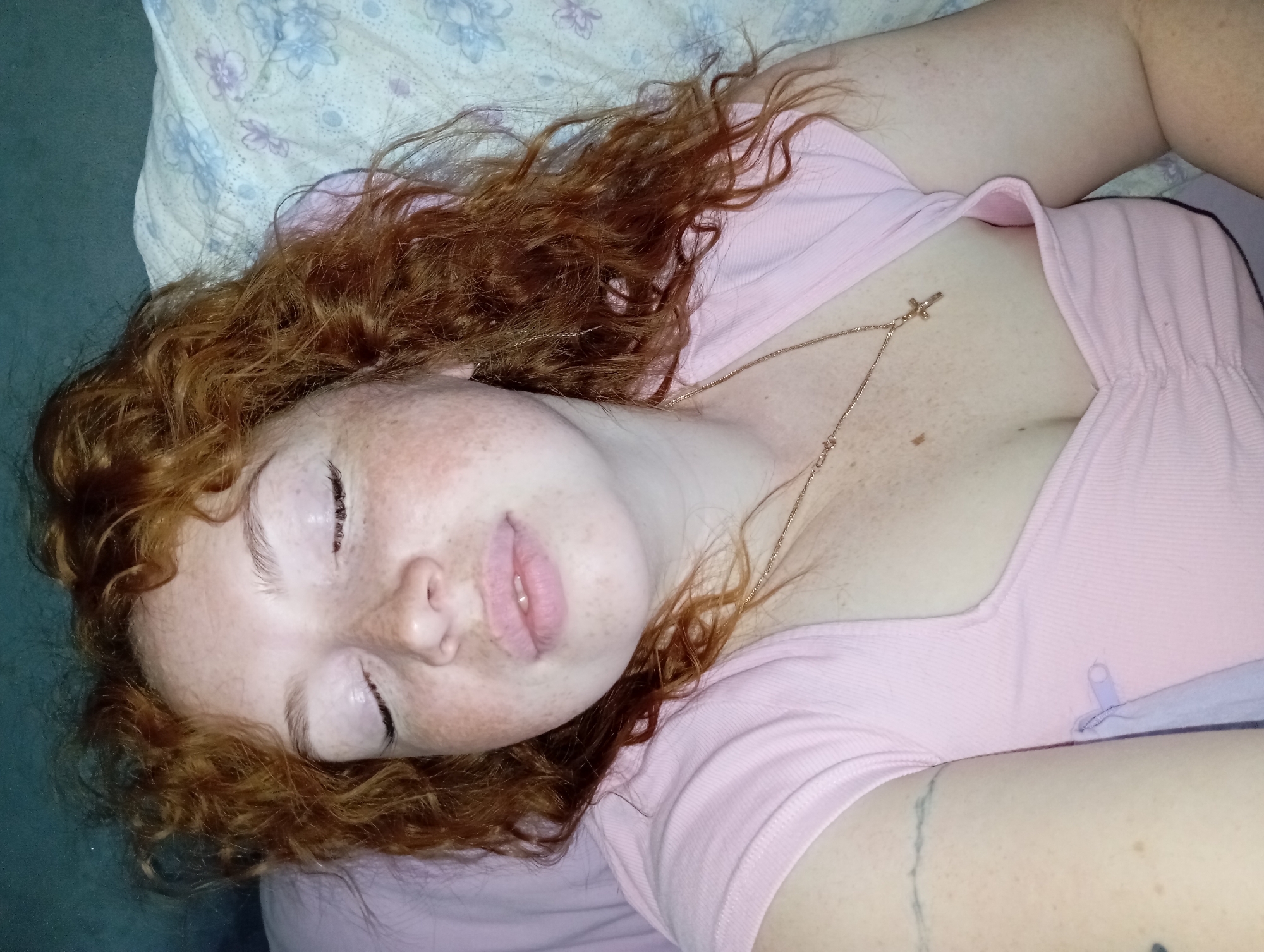 A fair-skinned woman with red curly hair and freckles lies on a bed. She wears a light pink top and a gold necklace with a cross pendant. Her eyes are closed and her face is facing the camera. The bedding has a floral pattern. There is a tattoo visible on her left arm. The environment is not possible to determine beyond the bed. The location of the image is unknown, and therefore cannot be placed in Yurgamysh, Russia.