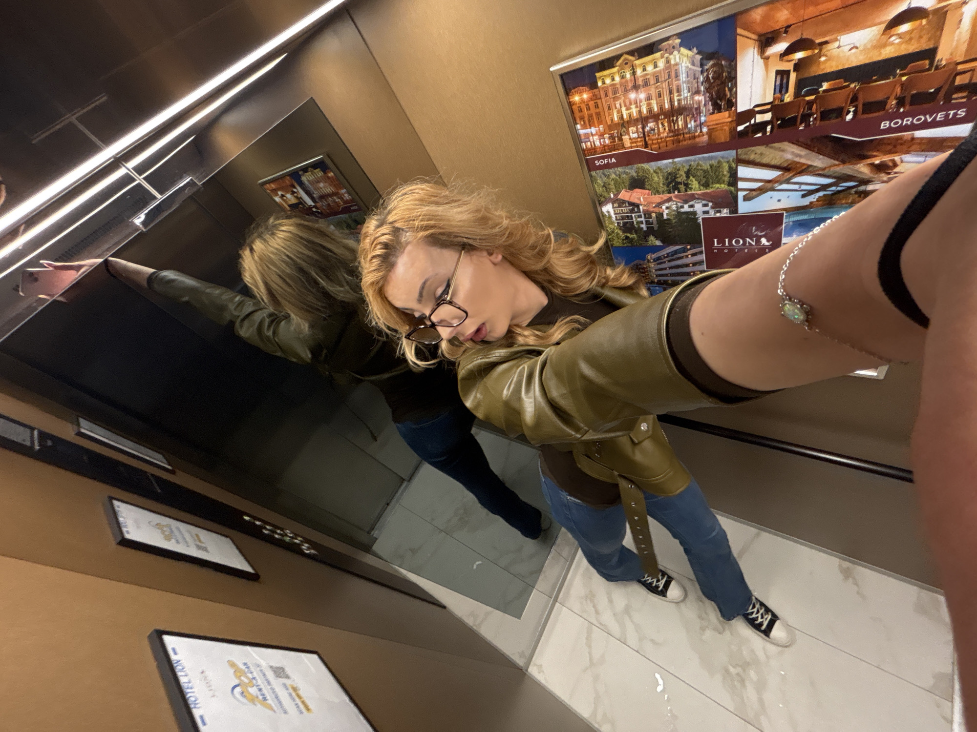 A female-presenting person with blonde hair, glasses, and a green jacket takes a selfie in an elevator. The elevator has posters of Sofia and Borovets, indicating the location is likely in Sofia, Bulgaria. The person wears blue jeans and black and white sneakers. The elevator has a mirror reflecting the person and the elevator interior. The floor is tiled.