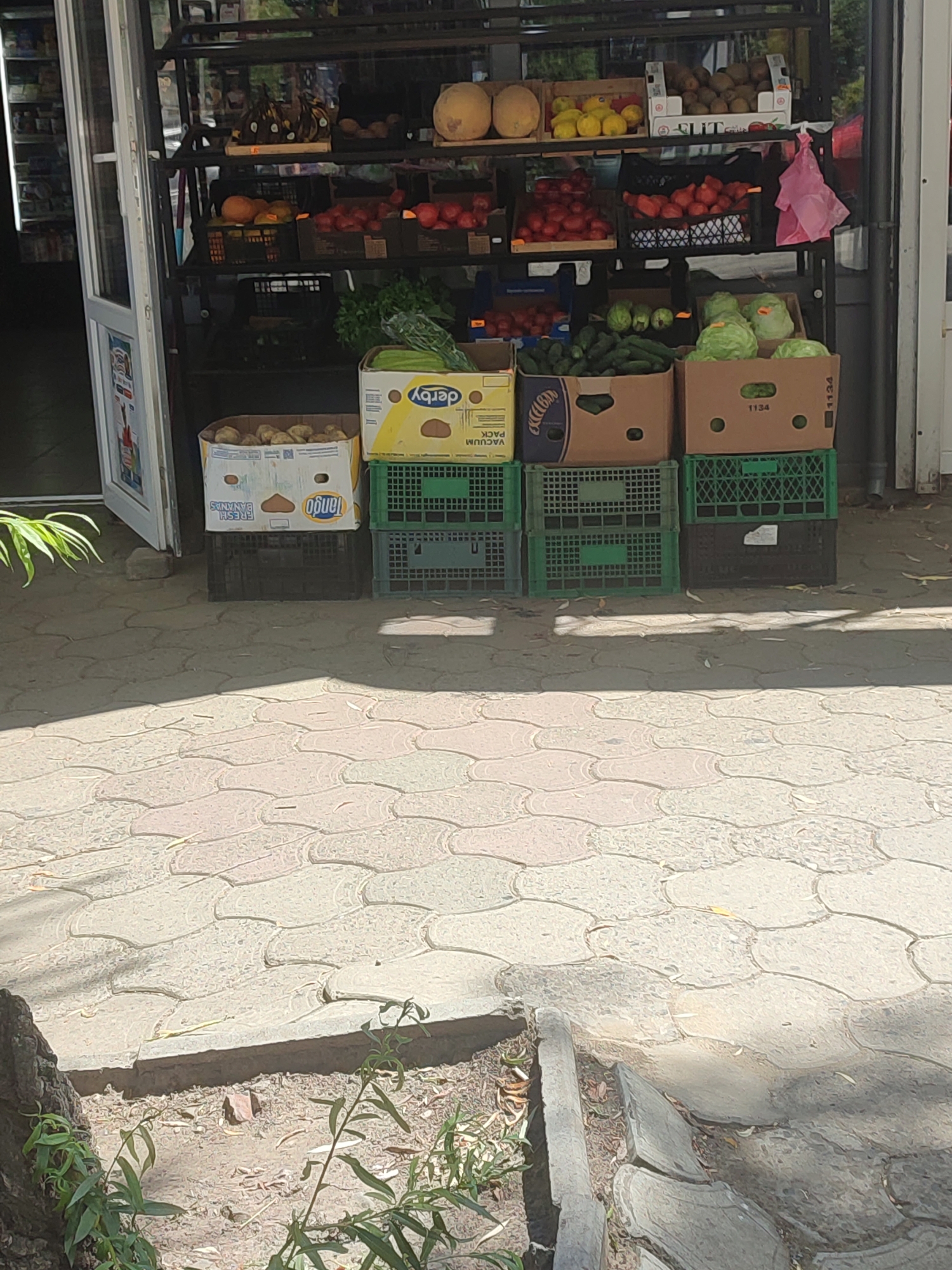 The image shows an outdoor fruit and vegetable stand in Chernivtsi, Ukraine. The stand is stocked with various produce including melons, lemons, tomatoes, cucumbers, potatoes, and lettuce. The items are displayed in cardboard boxes and plastic crates. The stand itself appears to be a metal shelving unit. The ground is paved with interlocking stone tiles. The lighting suggests it is daytime with some areas in shadow. There are no people visible in the image. The environment appears to be an urban setting, possibly a street or market area.