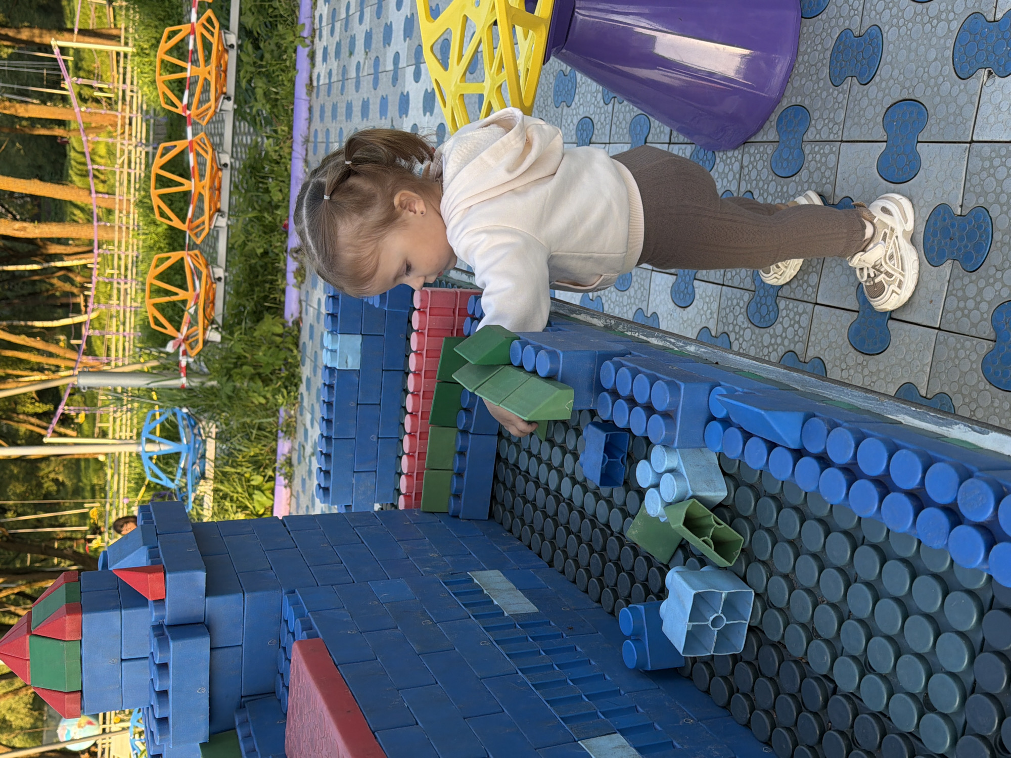 A young girl is playing with oversized building blocks at a playground, likely located in Sin'kovo, Russia. She is wearing a light-colored hoodie and brown pants, along with white sneakers. She is bent over, placing a green block onto a structure made of blue and red blocks. The blocks appear to be large and designed for children to build with. The playground has a soft, patterned tile surface and features other colorful play structures in the background, including yellow geometric shapes and a purple cone. The location appears to be outdoors with trees visible in the background, suggesting it is a park or recreational area.