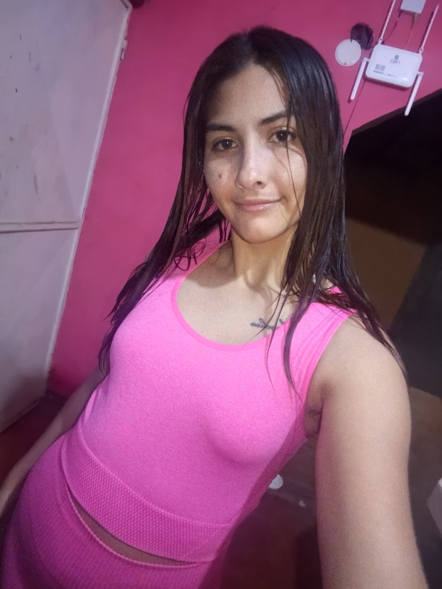 A young woman is captured in a selfie indoors, likely in Juan Griego, Venezuela. She has long, dark hair that appears damp or wet, falling over her shoulders. Her complexion is fair, and she sports a subtle, contented smile while looking directly at the camera. She is wearing a bright fuchsia or hot pink two-piece athletic outfit, consisting of a sleeveless top and a matching bottom (skirt or leggings) that features a ribbed or textured waistband, revealing a narrow strip of her midriff. A small, dark symbolic tattoo, possibly a cross or anchor, is visible on her upper left chest area.
The background features a vibrant fuchsia-pink wall. To the left, a portion of a white structure, possibly a wardrobe or cabinet, is visible. On the right side of the frame, mounted on the pink wall, is a white Wi-Fi router with two distinct antennas. Adjacent to the router, there's a circular white object, which could be a light fixture or another electronic device. Some unreadable text is faintly visible on the router. The lighting suggests an indoor setting, possibly under artificial light.