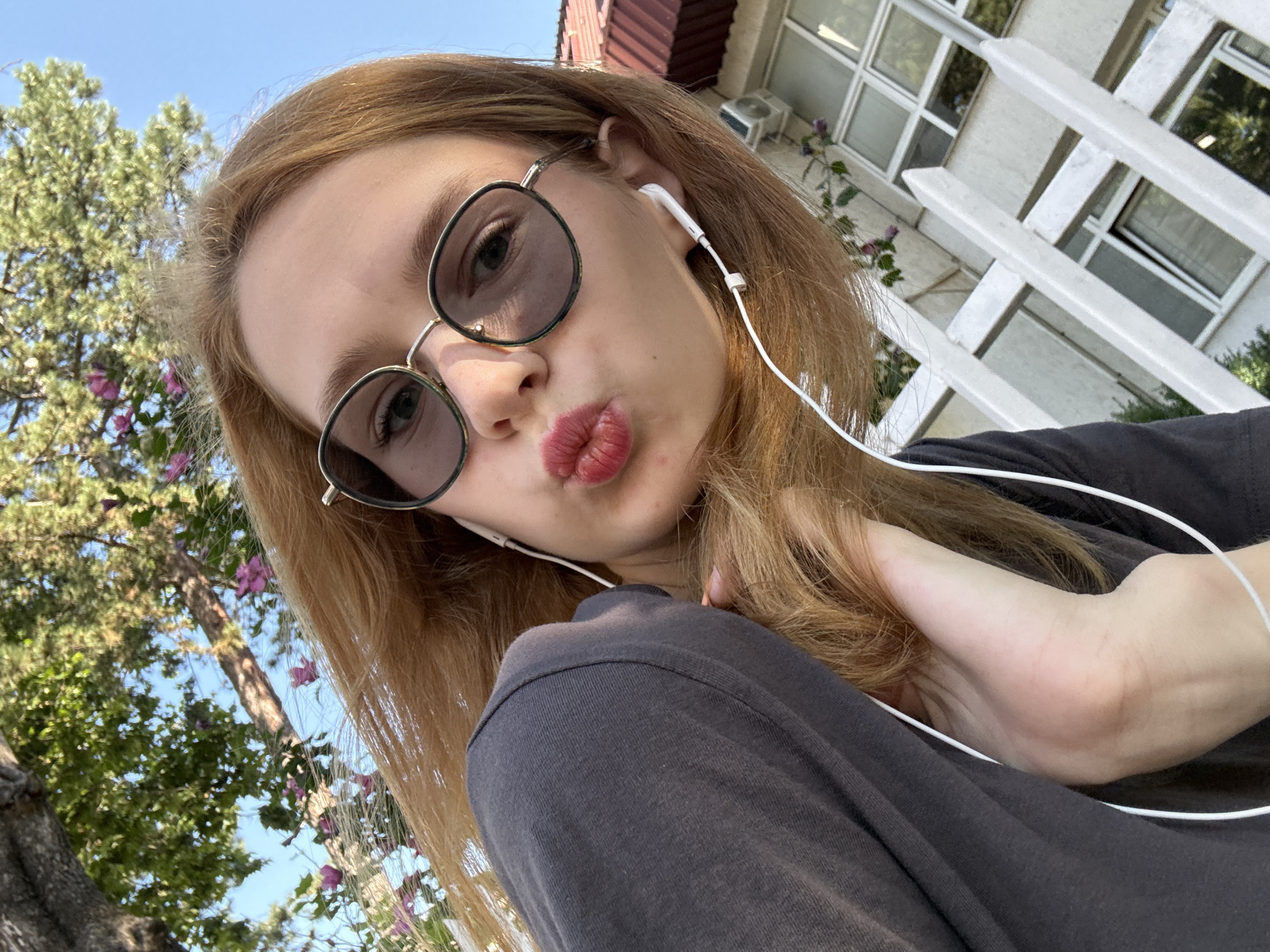 A young woman with long, reddish-blonde hair and sunglasses is taking a selfie outdoors. She is wearing a dark grey t-shirt and white earbuds. Her lips are pursed as if she is about to kiss. In the background, there are trees with green leaves and pink flowers, a blue sky, a white railing, and a building with a red roof and windows. The overall scene suggests a casual, sunny day. The location is identified as Kragujevac, Serbia.