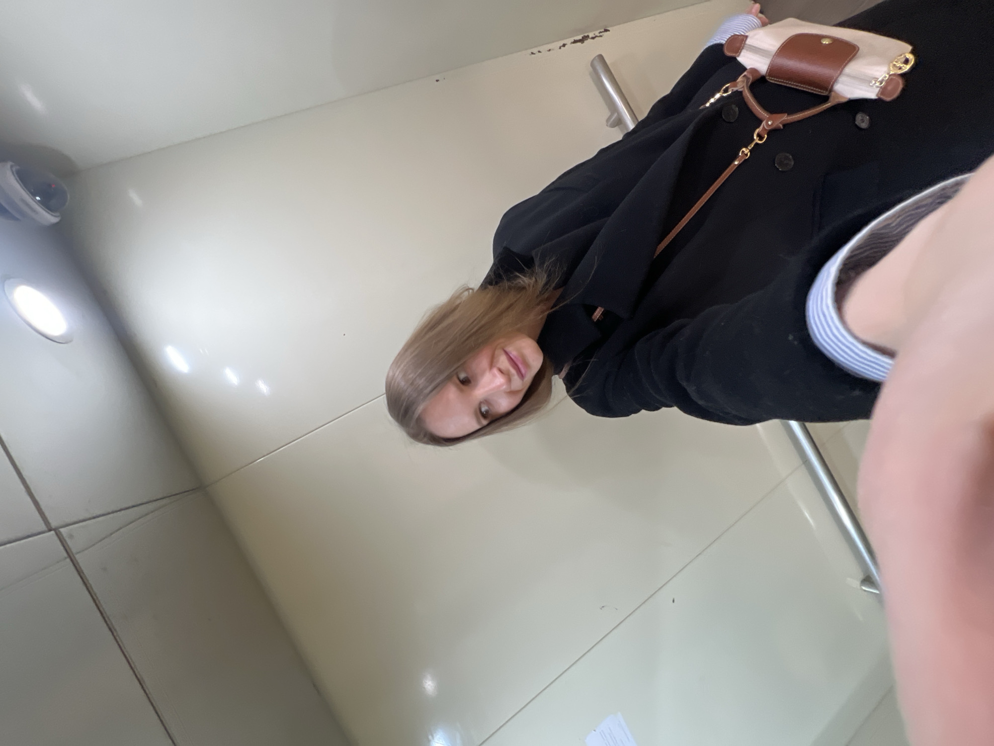 A person is in an enclosed space, possibly an elevator, as suggested by the safety bar and tiled walls. The space is brightly lit. The person has light brown hair and is wearing a black coat with visible buttons and a blue and white striped shirt underneath. A small tan purse with brown leather details hangs across their body. The exact location within Khimki, Russia, cannot be determined from the visual information.