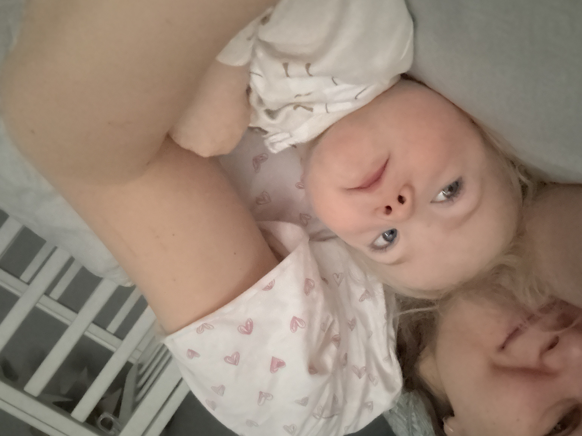 A close-up shot shows an adult and a baby lying down in a crib, both facing upwards towards the camera. The baby has fair skin, blonde hair, and blue eyes. They are wearing a white outfit with pink hearts printed on it. The adult has fair skin, blonde hair, and is positioned slightly behind the baby, with part of their face visible. The crib has white bars. The location is unknown.