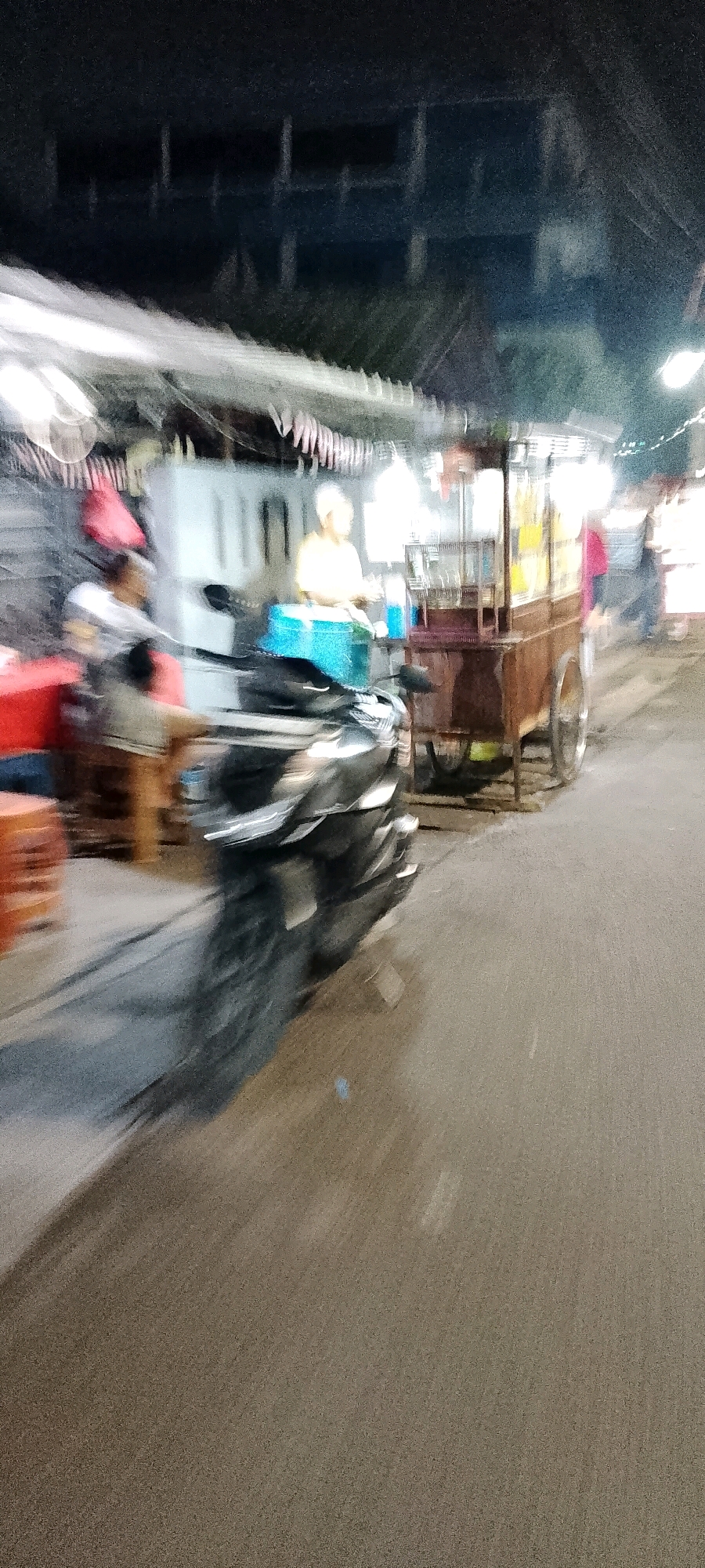 This is a blurry, night-time, street-level photograph taken in Bekasi, Indonesia. The scene depicts a bustling street food stall. In the foreground, a dark-colored motorcycle is partially visible, blurred by motion. Behind the motorcycle, a food cart with metal shelving and wheels is positioned, laden with goods and illuminated by overhead lights. A person wearing a light-colored top appears to be working at the stall. To the left of the food cart, a red object, possibly a table or seating, is visible. The background shows a building with a dark, partially visible facade and multiple vertical structures. The overall impression is of a lively, outdoor dining area with people and activity.