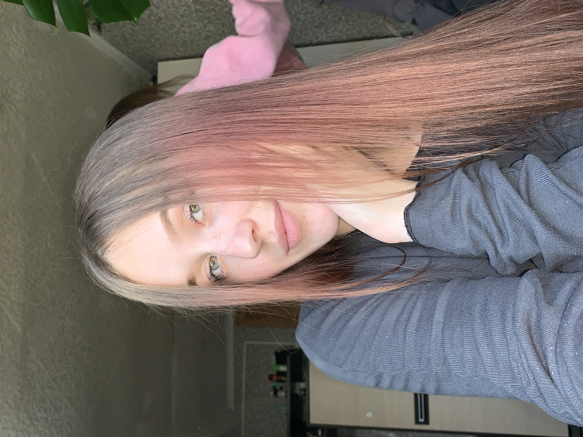 A close-up, slightly angled portrait shows a fair-skinned young woman with long, light brown hair transitioning to pink at the ends. Her eyes are green, and she has freckles on her nose. She wears a gray, textured, long-sleeved shirt. Her hand is visible near her neck. The background includes a blurred view of a room with light-colored walls, a plant in the upper left corner, and a glimpse of a shelving unit. The location is specified as the city of Фоки, Russia. No actions or interactions are evident.