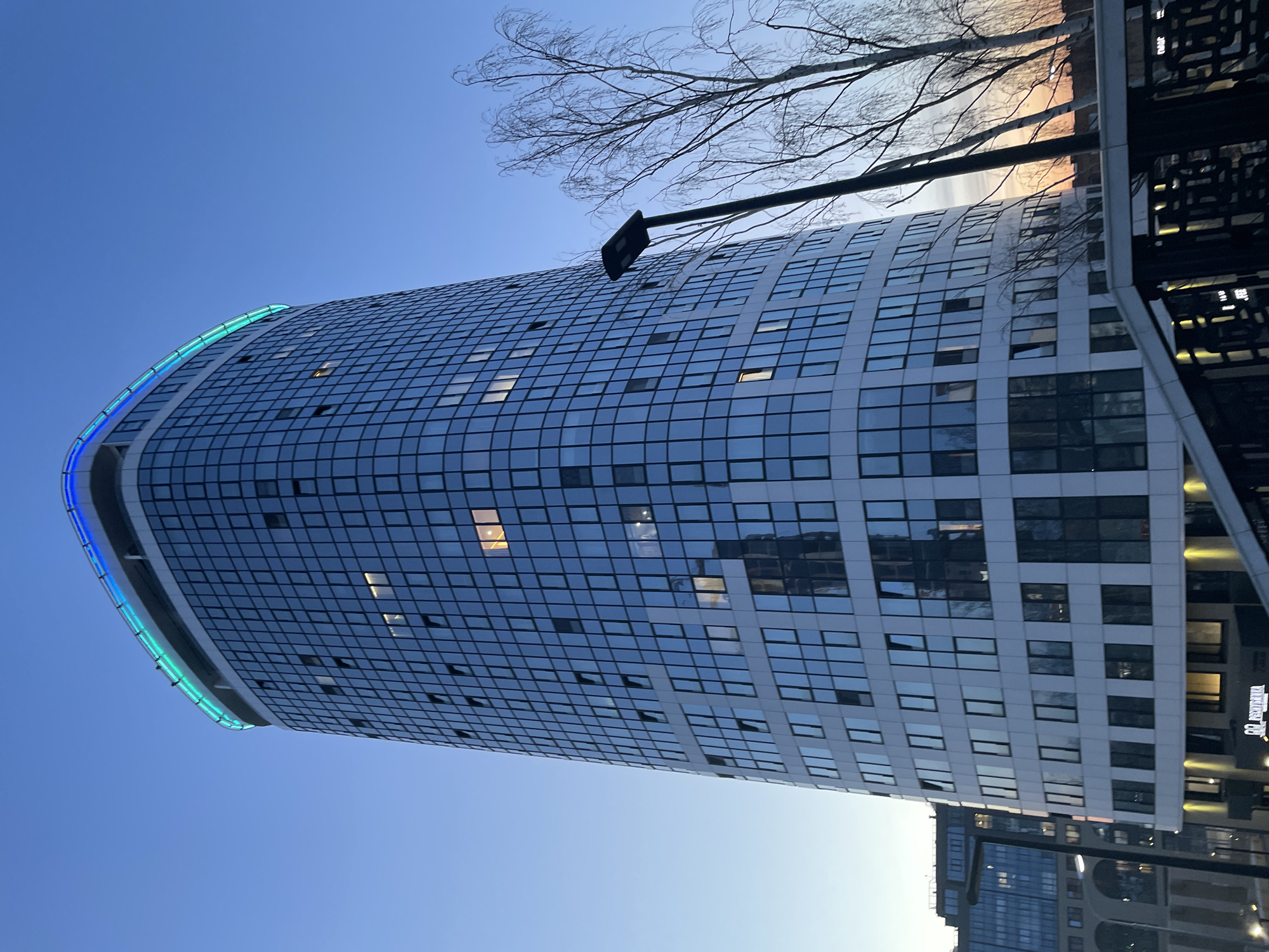 A tall, cylindrical building dominates the scene, situated in Izhevsk, Russia. The building has a grid-like facade comprised of numerous windows, some of which have lights on inside. The top of the building is adorned with a blue and green light strip. A few bare tree branches are visible near the top right corner. In the foreground on the right, there's a section of a metal structure. The sky is blue.