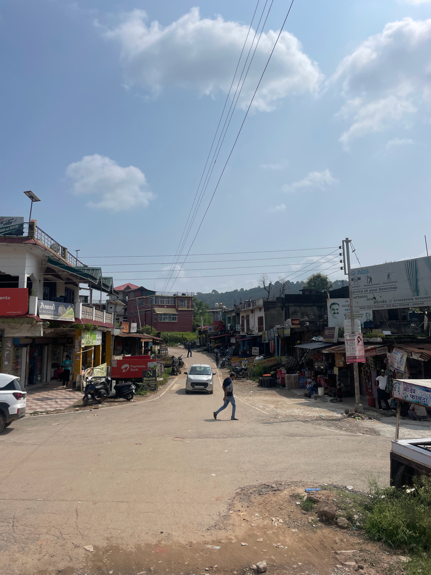 The image depicts a street scene in Basa Balhu, India, under a bright, cloudy sky. The road, paved with asphalt, shows signs of wear with some potholes and debris scattered on the surface. On the left, a white SUV is parked, and a person is walking across the street. Adjacent to the road are buildings, some with balconies and awnings, housing various shops and businesses. Signs for "Amul" and "Payments Bank" are visible. Several motorcycles are parked along the side of the road. To the right, more shops and stalls line the street, with people visible in and around them. A large billboard with graphics and text is mounted on a pole. Further down the road, more buildings and trees are seen, with hills in the background. The overall atmosphere suggests a bustling, everyday street in a small town or city.