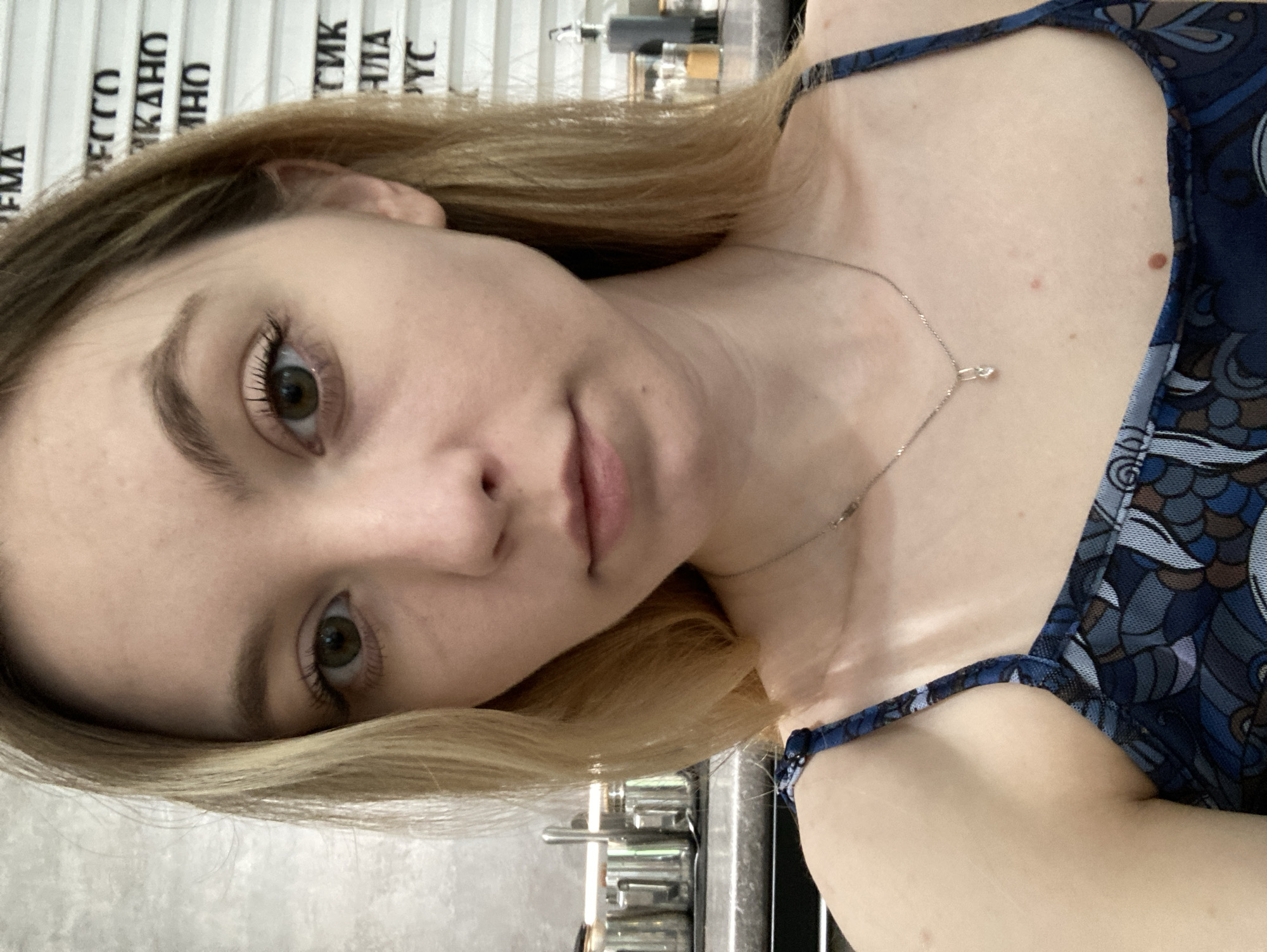 A young woman with light brown hair and green eyes is posing for a selfie. She is wearing a dark blue patterned sleeveless top and a delicate silver necklace. The background appears to be a cafe or restaurant, judging by the menu board with Russian writing and the coffee machine visible behind her. It is implied that the location is Chaikovsky, Russia.