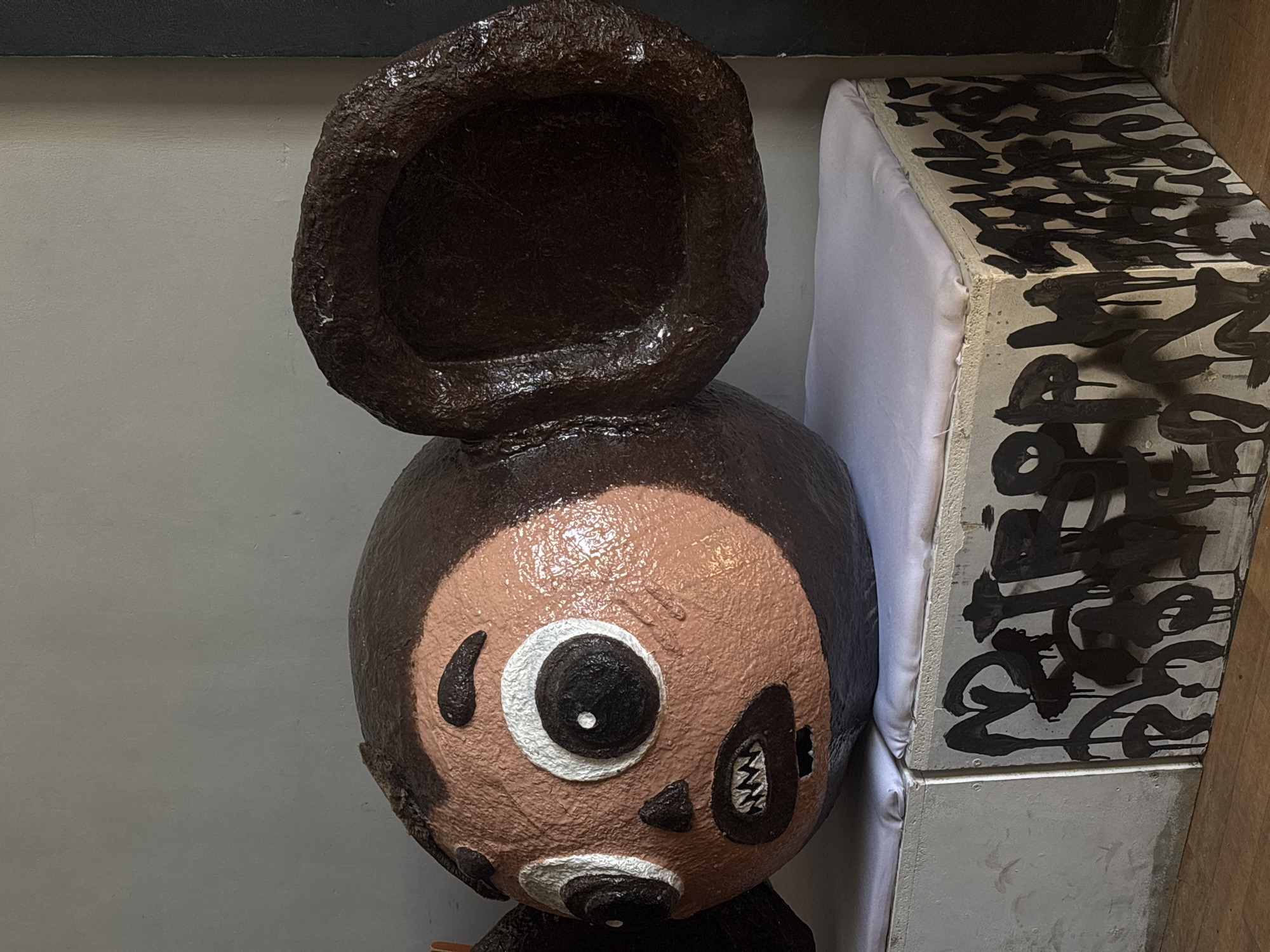 A close-up shows a folk art sculpture of a character with a stylized, expressive face, possibly inspired by traditional Russian folklore. The sculpture is positioned near a painted and upholstered box in what appears to be an indoor setting in Perm, Russia. The face features exaggerated eyes and mouth, rendered in a somewhat crude style with a contrasting color palette. The background is primarily a neutral wall color, and the overall lighting is soft and diffused, highlighting the textured surface of the sculpture.