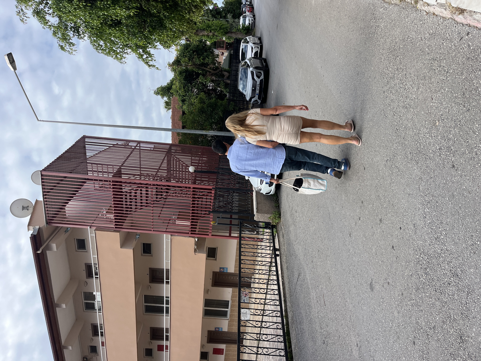 A couple walks along a street in Alanya, Turkey, with a beige building to their left and parked cars ahead. The man wears a blue shirt, dark pants, and blue shoes, carrying a white bag. The woman has blonde hair and wears a light-colored dress with sandals. A black wrought-iron fence runs along the side of the building, which has a maroon-colored roof structure. A street light stands tall next to the building, with green trees visible above and along the street. The sky is overcast.
