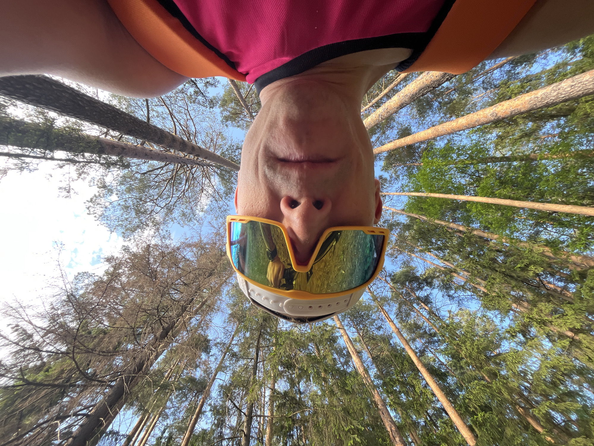 The image shows a person lying on their back in a forest, looking up at the trees. The person is wearing sunglasses with reflective lenses, a white hat, and an orange and pink shirt. The trees are tall and thin, and the sky is visible through the branches. The image was taken in Chaikovsky, Russia.