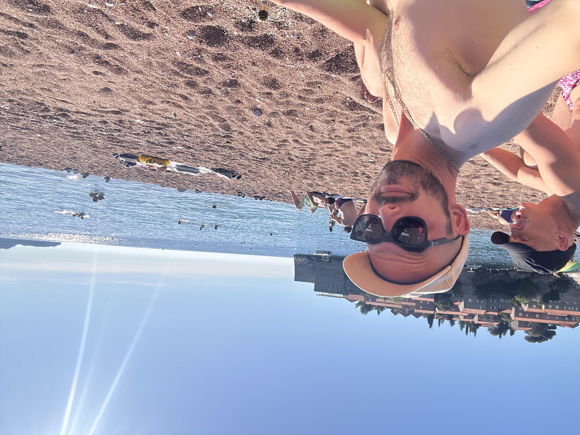 This is a vertical, upside-down selfie taken on a beach. The foreground is dominated by the faces of two men. The man on the left is wearing sunglasses and a light-colored baseball cap, with a chain necklace visible. The man on the right is wearing a dark baseball cap. The background shows a pebble beach transitioning into the clear blue ocean. Several people are visible in the water, some swimming or wading. In the distance, across the water, is a picturesque island settlement with red-tiled roofs, consistent with the location of Sveti Stefan, Montenegro. The sky is a bright, clear blue, suggesting a sunny day, likely in the late morning or early afternoon given the sun's intensity and the angle of shadows. The overall mood appears relaxed and summery. No discernible text is visible in the image.