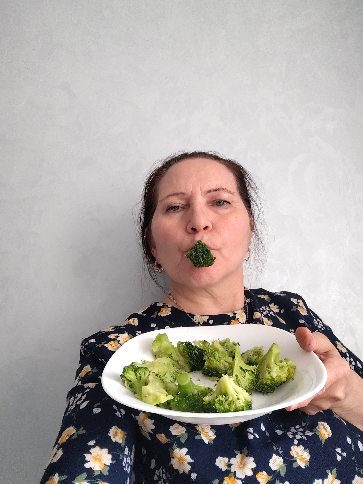 A woman with brown hair and light skin holds a white oval plate filled with cooked broccoli. She has a piece of broccoli in her mouth. The woman is wearing a dark blue dress with a yellow floral pattern, gold hoop earrings, and a necklace. The background is a plain, light gray wall. The photograph was taken in the city of Sarapul, Russia.