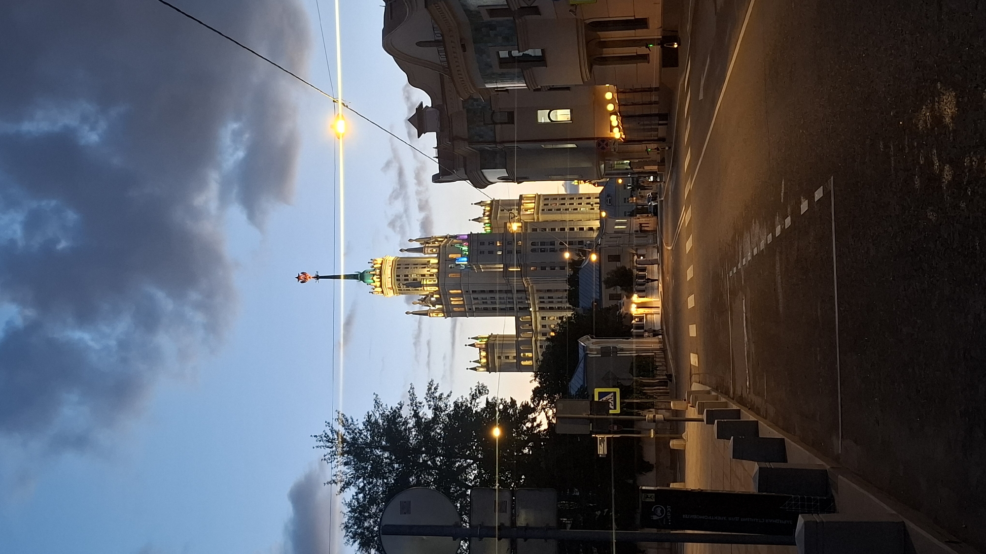 A vertical, eye-level shot captures the Kotelnicheskaya Embankment Building in Moscow at dusk. The imposing Stalinist skyscraper, illuminated with warm yellow lights, dominates the skyline. Its spire is topped with a red star. The sky above is a mix of deep blue and dark, swirling clouds. In the foreground, a street with white dashed lines and crosswalks runs towards the building. The streetlights cast a warm glow on the wet pavement, suggesting recent rain. On the right, other buildings with illuminated windows and ornate facades line the street. On the left, a sidewalk with bollards and a sign for an electric vehicle charging station is visible. A silhouette of a tree is on the far left. No people are clearly visible.