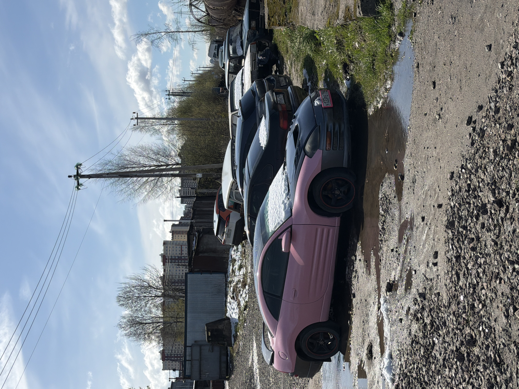 The image captures an outdoor scene in Lavriki, Russia, depicting a row of parked cars on a gravel and dirt lot. The perspective is from slightly below, looking along the line of vehicles. In the immediate foreground, a striking pink and black two-door coupe, possibly a Mitsubishi Eclipse, is prominently displayed, its hood and roof partially covered in patches of snow. A Russian license plate, "У 433РЕ 198," is visible on its front bumper.
Behind the pink car, several other older-model cars, mostly dark-colored sedans and coupes, are parked in succession, also bearing traces of snow on their surfaces. The ground beneath and around the cars is a rough mix of gravel, dirt, and mud, interspersed with numerous puddles of water that reflect the bright sky. A strip of green grass and sparse, bare-branched trees runs along the edge of the lot, separating it from the line of utility poles and overhead power lines that stretch across the mid-ground.
In the distance, multi-story apartment buildings are visible under a bright, partly cloudy sky, featuring broad streaks of white clouds against a vibrant blue. The presence of both snow and significant puddles suggests that the image was taken during early spring, likely during a period of thaw. The time of day is clearly daylight, with shadows indicating a sunlit scene. No people are present, and there is no specific activity taking place; the cars appear stationary, lending a quiet and somewhat desolate atmosphere to the scene.