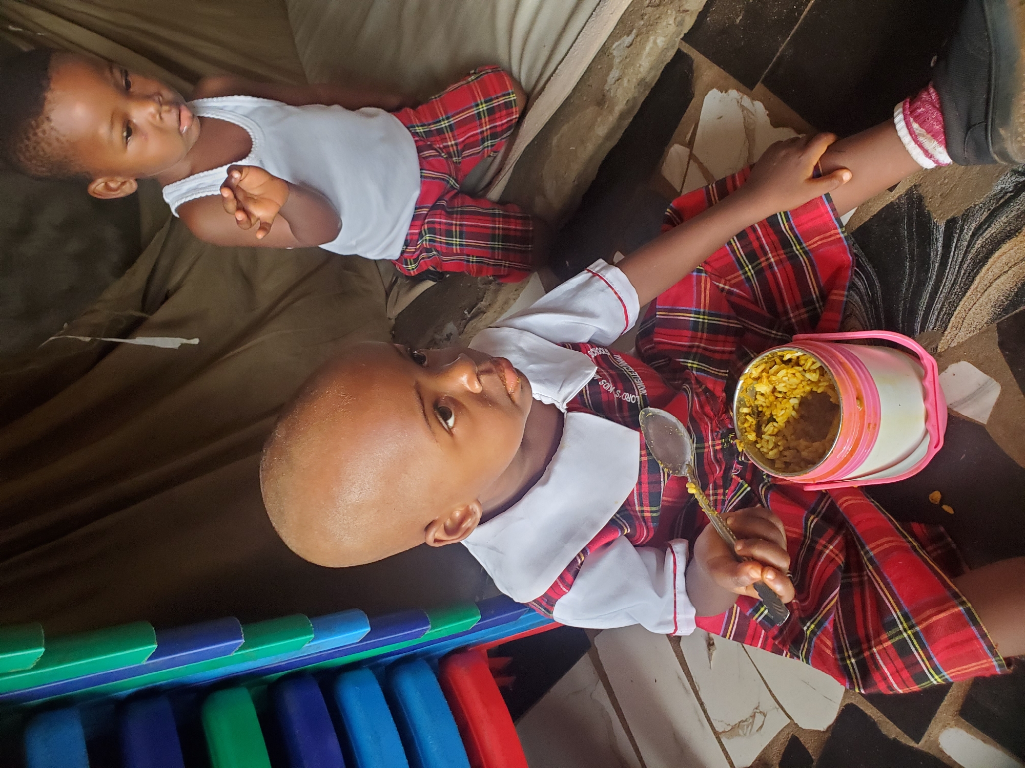 Two children are present in an interior setting. One child is seated on the floor eating from a container with a spoon; the container appears to be a pink and white thermos with a visible yellow food substance. The child is wearing a red and black plaid dress-like garment over a white collared shirt. The second child is lying on a tan surface. This child is dressed in a white sleeveless shirt and red and black plaid pants. The location is indicated as Ukwu-Abwa, Nigeria; further environmental details include a multi-colored stack of plastic chairs and a tiled floor pattern.