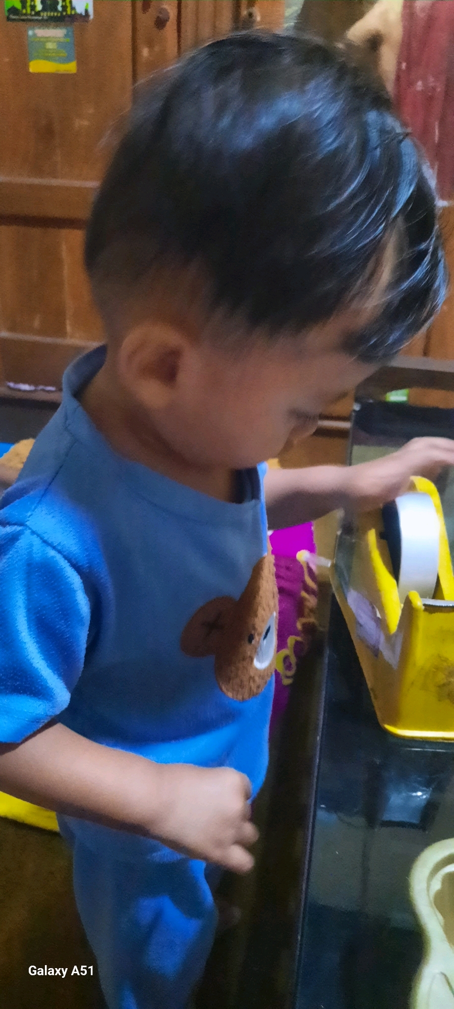 A young child, facing away from the camera and looking down at a yellow tape dispenser, is visible in this image. The child is wearing a blue shirt with a brown bear graphic on the chest. The setting appears to be indoors, possibly a home, with wooden furniture and a dark countertop. The lower-left corner of the image displays the text "Galaxy A51", indicating the device used for capture. The location is specified as Ploso, Indonesia.