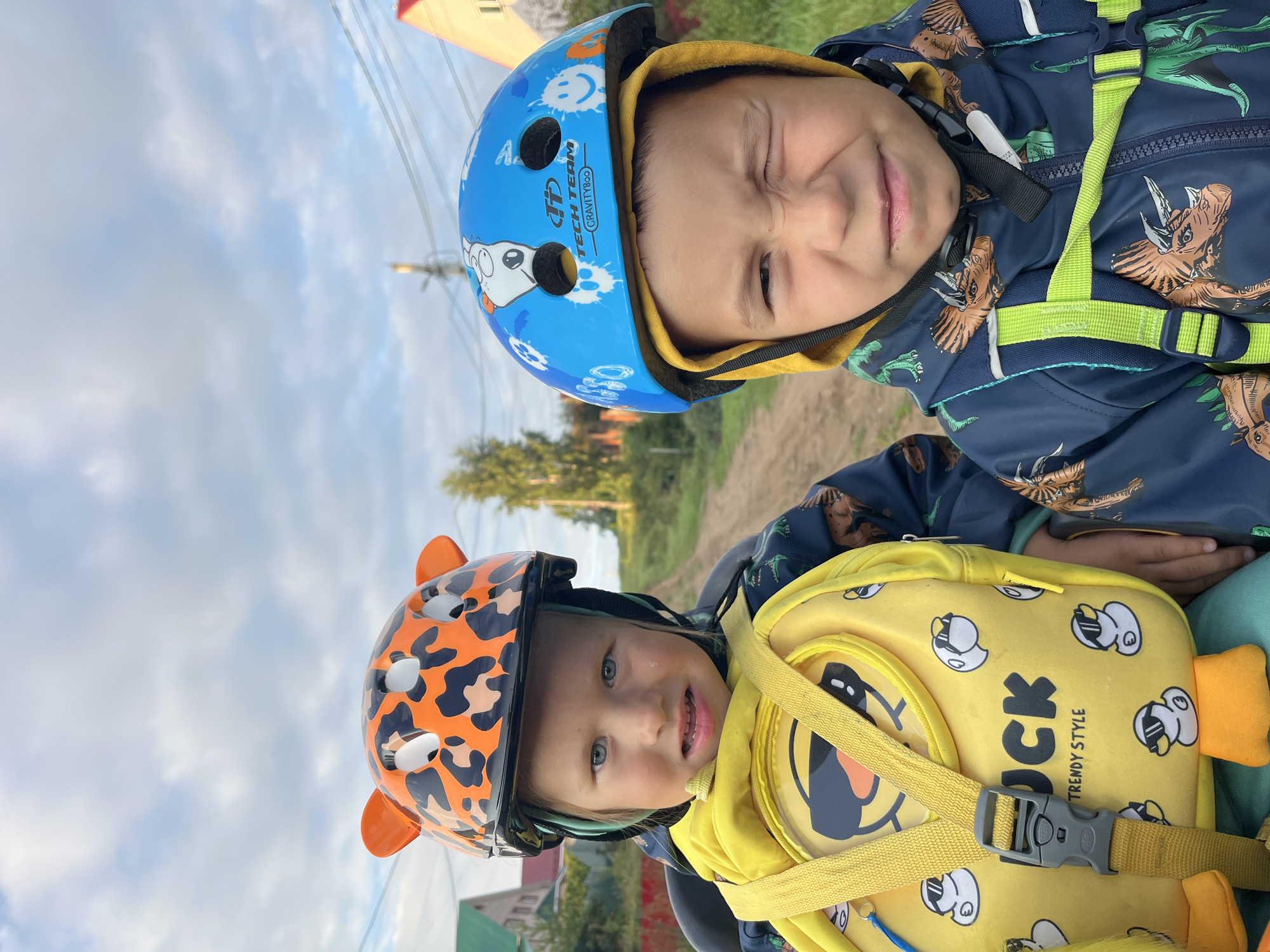 Two young boys are seated in what appears to be a bicycle seat or stroller, facing the camera. The boy on the left wears an orange helmet with a leopard print design and large ears, and a yellow backpack with cartoon ducks. He has blue eyes and is looking directly at the camera. The boy on the right wears a blue helmet with various cartoon characters and text that appears to say "TECH TEAM GRAVITY". He is winking at the camera. Both boys are wearing jackets with dinosaur prints. The background suggests a rural or suburban setting with a dirt road, trees, and houses in the distance. The sky is cloudy. The location is identified as Chaikovsky, Russia.