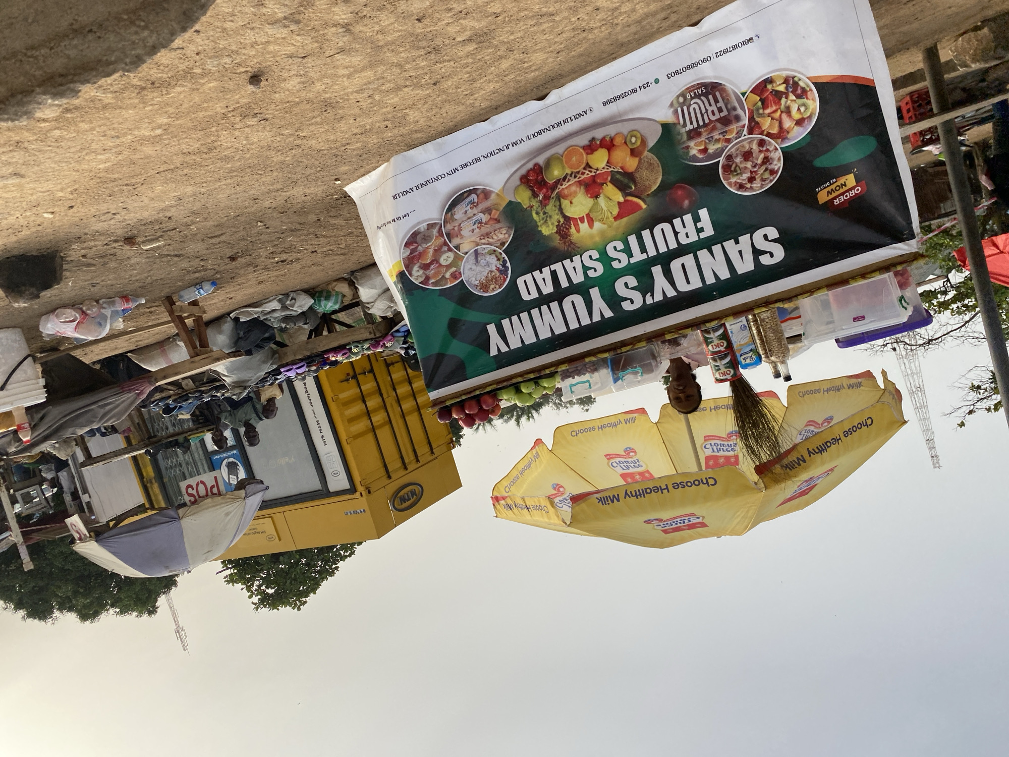 This outdoor image captures a vibrant market scene in Bukuru, Nigeria, under bright, clear daylight. The primary subject is a roadside fruit salad stand featuring a large, colorful banner for "SANDY'S YUMMY FRUITS SALAD." The banner displays appetizing images of fruit salads, the slogan "ORDER NOW WE DELIVER," and contact information including phone numbers (+234 8102568398, 8101871922, 09068807803) and an address: "ANGULDI ROUNABOUT/ VOM JUNCTION, BEFORE MIN CONTAINER ANGULDL." Various items like packaged nuts, apples, and plastic containers are visible on the stall's counter.
To the right, a yellow umbrella provides shade, emblazoned with "Choose Healthy Milk" and logos for "Peak" and "Oki" dairy products. A person's head is partially visible beneath it. In the background, a large yellow shipping container branded with "MTN" serves as a mobile network service point, displaying "Register your MTN SIM," "Y'ello," and a prominent "POS" (Point of Sale) sign. Two men are standing near this container. A smaller, grey and white umbrella is also seen near the MTN container.
The ground is unpaved and dusty, with scattered debris including plastic bottles. Sparse trees and a communication mast are visible in the background against a bright sky. The scene depicts a typical informal street vending operation, likely offering fresh food and essential services.