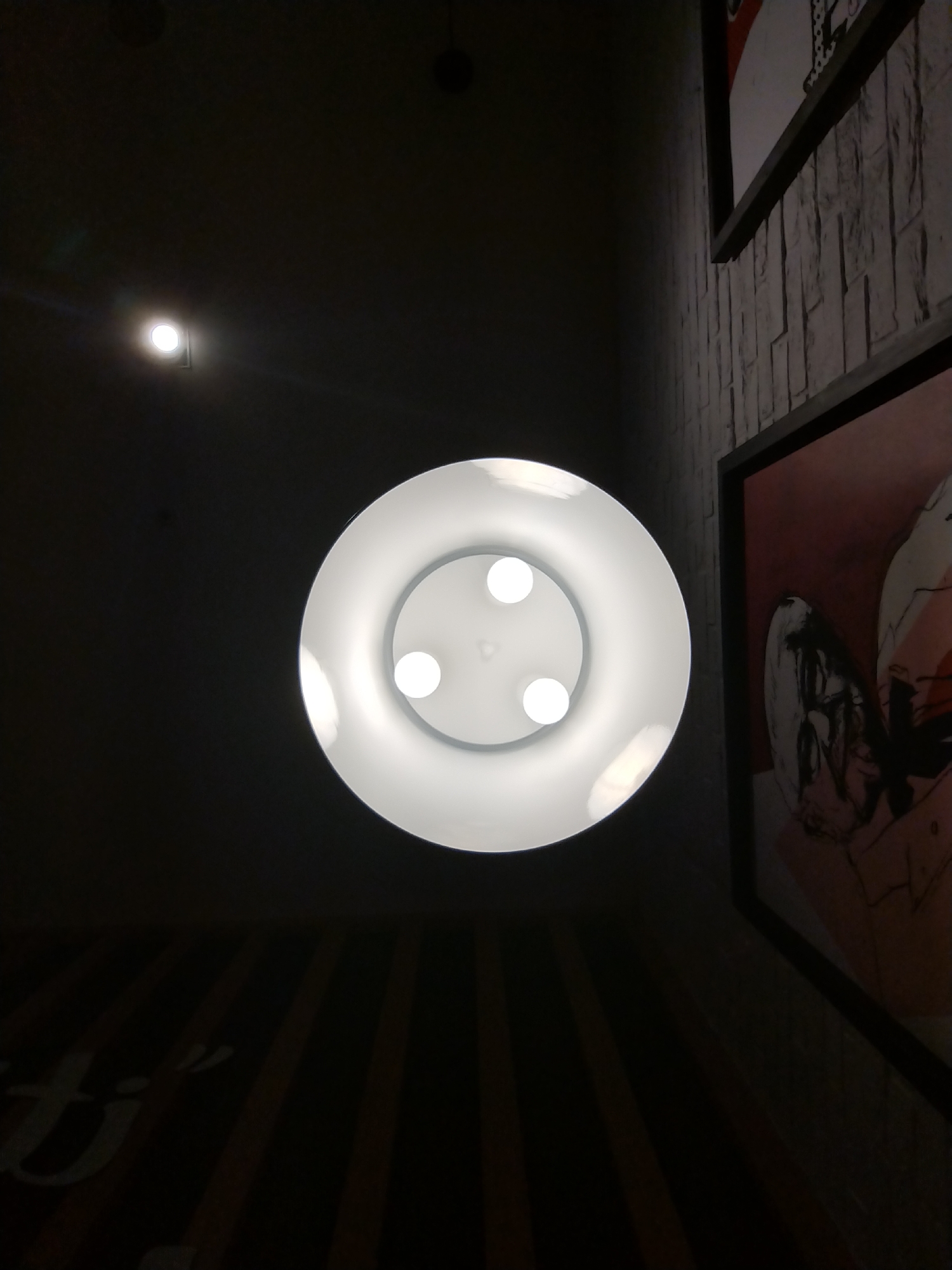 The image presents an upward-angled view of an indoor space, dominated by artificial light sources. A large, circular ceiling lamp with a white reflector is centrally placed, housing three bright, circular bulbs that emit a strong white light. To the upper left, a smaller, square-shaped recessed light fixture also illuminates the dark ceiling.
On the right side of the frame, a wall with a light-colored, textured surface resembling stacked bricks is visible. This wall features a framed artwork or poster, partially seen, which appears to depict abstract or illustrative elements in tones of red, black, and white. Below the central lamp, extending into the bottom left corner, is a dark surface with vertical stripes. Upon this striped surface, light-colored, stylized text is partially visible, showing "ti'" followed by an apostrophe, suggesting it is part of a larger word.
The overall environment is dimly lit, with the bright lights creating significant contrast against the dark background and accentuating the illuminated architectural features and decor. The scene captures the ambiance of a commercial establishment, such as a cafe, restaurant, or bar, possibly in Varna, Bulgaria, given the location context. No people are present, and the image focuses on the interior lighting and design elements.