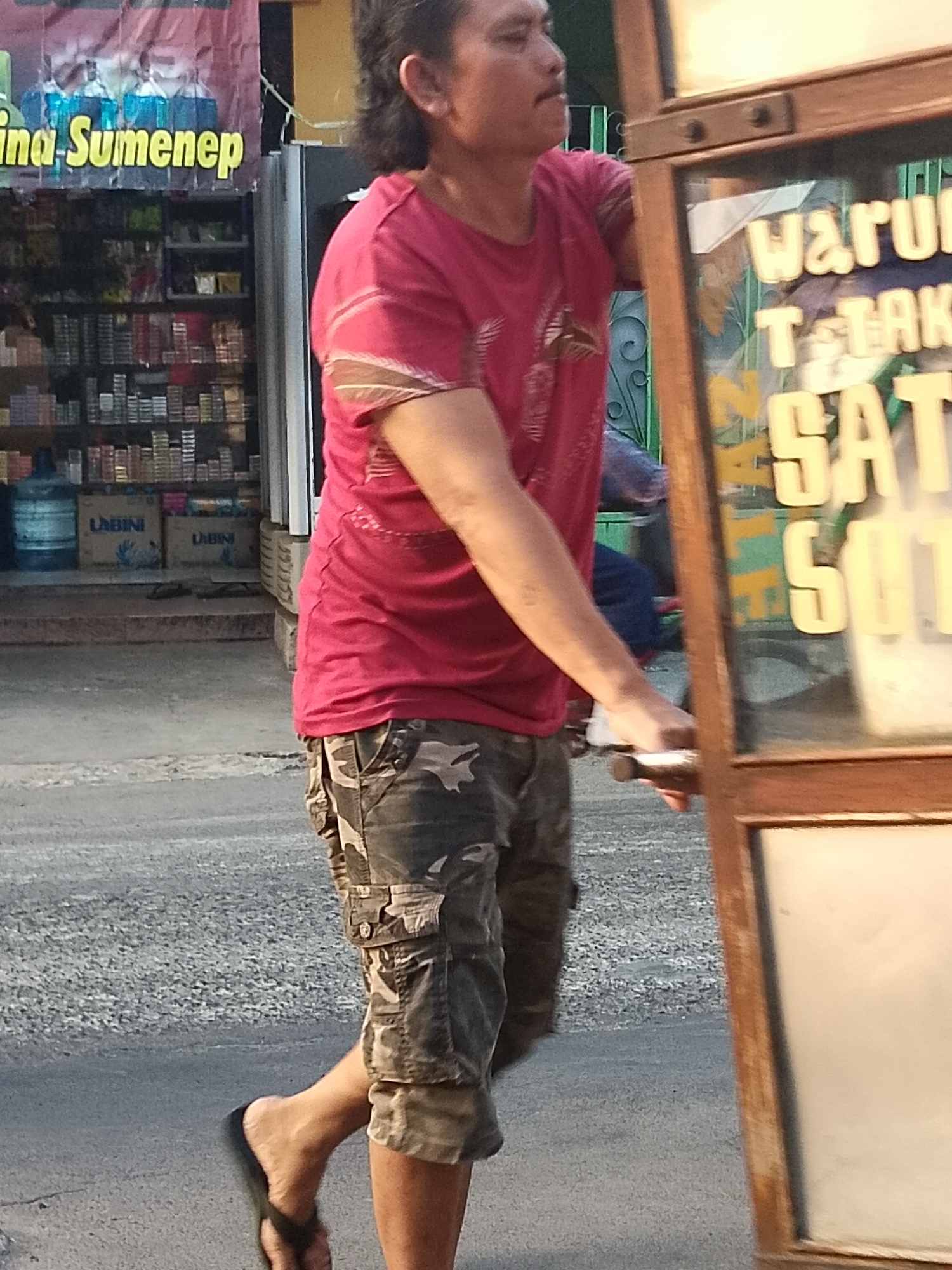 A man is seen from the chest down, wearing a red t-shirt and camouflage cargo shorts, walking along an asphalt road. He is pushing a wooden cart with a glass display window that has text printed on it. The background shows a shop with shelves stocked with items, including bottled drinks and boxes labeled "LABINI." The text on the cart's window appears to read "WARUNG T-TAK SATU", suggesting it might be a small food or drink stall. The overall scene depicts a street-level view in what appears to be a commercial area in Surabaya, Indonesia.