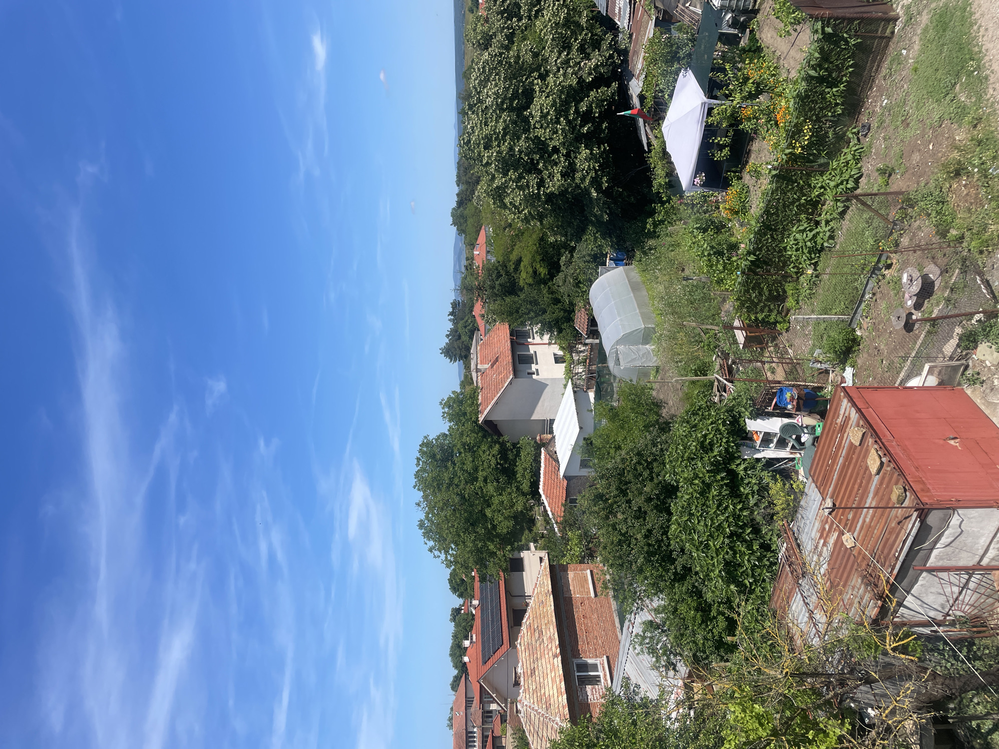 The image captures a view of a residential area in Debelt, Bulgaria, under a clear blue sky with scattered wispy clouds. In the foreground, a red metal shed with a rusty corrugated metal roof is visible. To the left and extending towards the center, various garden plots with fencing, plants, and some structures are present, including a white canopy tent and a greenhouse. A Bulgarian flag is attached to a pole near the canopy tent.
Further back, a collection of houses with terracotta tile roofs and some with modern solar panels are interspersed with lush green trees. The houses appear to be constructed from brick and stucco. The terrain slopes slightly, suggesting a suburban or rural setting within the town. The overall impression is one of a lived-in, somewhat rustic environment on a bright, sunny day. There are no people visible in the image.