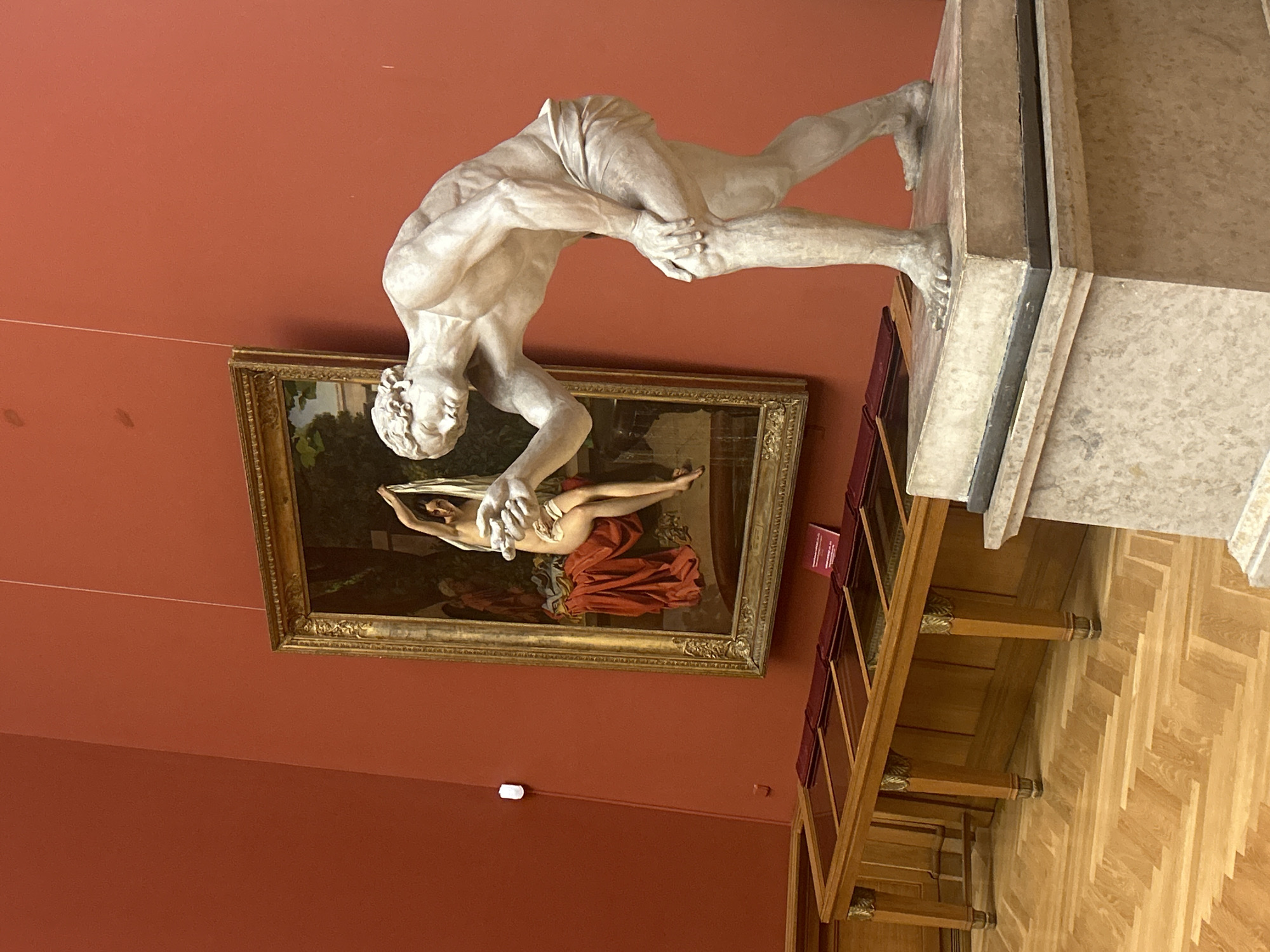 This image depicts a museum interior with art on display. A classical marble statue of a male figure, leaning forward with one hand on his knee, is positioned in the foreground, to the right of the frame. Behind the statue, a framed painting hangs on a reddish-orange wall. The painting shows a nude female figure with dark hair, holding a white cloth. She is seated on a draped red fabric next to a green leafy background. To the left of the statue and painting, there is a long wooden display case with a glass top. The floor is a polished parquet in a herringbone pattern. The lighting in the room appears to be artificial, with a visible light fixture on the wall in the background. The scene is likely within the State Hermitage Museum in Saint Petersburg, Russia, given the typical presentation of art in such institutions.