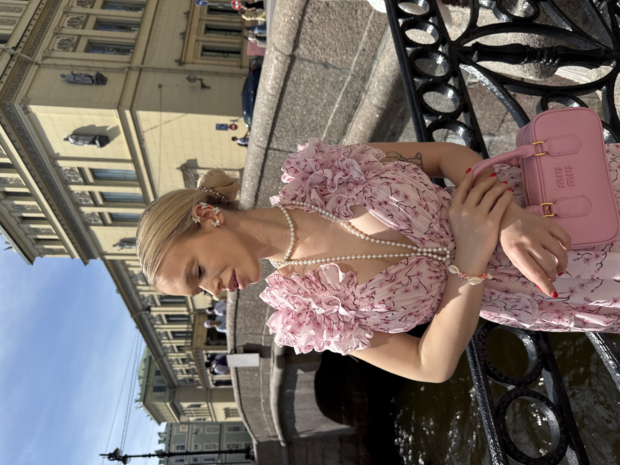 A young Caucasian woman with blonde hair pulled into a bun is standing on a bridge in Saint Petersburg, Russia. She is wearing a pink floral dress with ruffled sleeves, a pearl necklace, a shell bracelet, and butterfly-shaped earrings. She is holding a small pink handbag. The woman is looking to her right, with her eyes closed and her head tilted slightly. In the background, there is a canal with a stone bridge arching over it. Further back, there are historic buildings with classical architecture, typical of Saint Petersburg. The sky is blue with scattered clouds, and the sun is shining.