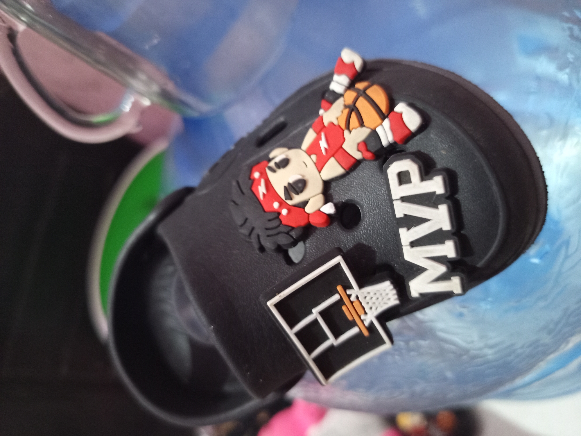 This is a close-up, eye-level, outdoor shot of a black slide-style shoe decorated with a cartoon basketball player charm and a miniature basketball hoop charm, along with the letters "MVP" in white. The shoe is resting on a textured, light blue surface, possibly a cooler or water container, which appears to be outdoors. In the background, out of focus, are colorful objects, one of which is a bright pink plush toy. The lighting is bright and casts shadows. The image does not contain any recognizable landmarks or contextual information that would indicate its location within the city of Tebel, Indonesia.