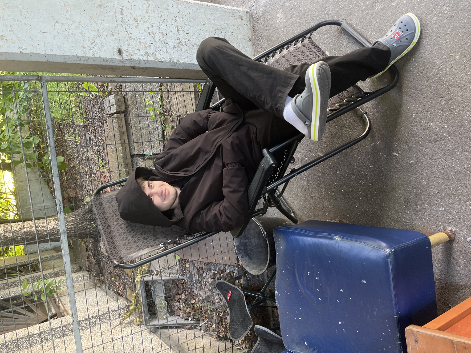 A young person is sitting in a black reclining chair on a concrete surface. They are wearing a dark hooded jacket, dark pants, white socks, and grey Crocs with a lime green stripe. The person is smiling and looking at the camera. To the left of the chair, there is a blue ottoman with wooden legs and some dirt on its surface. Behind the chair, there is a metal fence and some trees and vegetation. To the right of the chair is a white column. The scene appears to be outdoors, possibly in a backyard or a courtyard. The presence of the fence and the style of architecture suggest a location in Saint Petersburg, Russia.