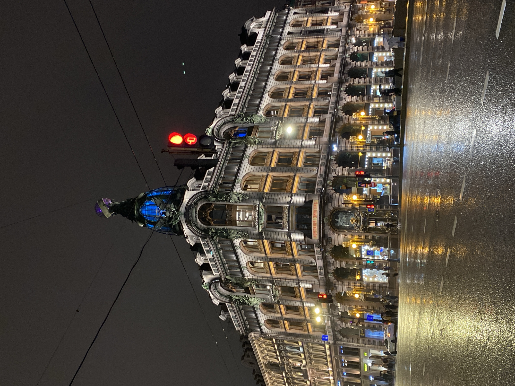 The image depicts a night scene in Saint Petersburg, Russia, featuring the iconic Eliseyev Emporium building. The building is ornately decorated, with multiple floors of illuminated windows and sculptural elements. The building's dome is lit up with blue light. Adjacent to the building is a wet street reflecting the city lights. People can be seen walking along the sidewalk near the building. Streetlights and traffic signals are visible, indicating urban surroundings. The sky is dark, suggesting it is late in the evening.