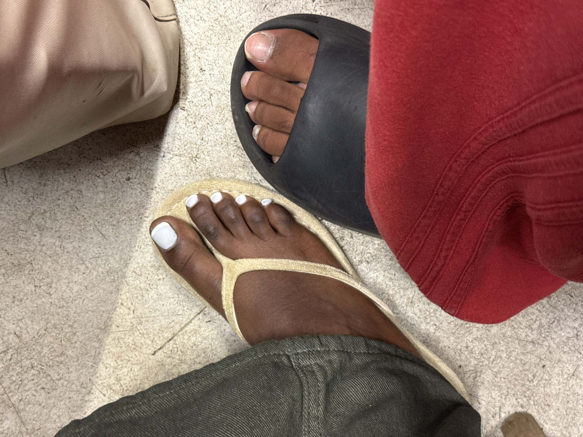 The image depicts two pairs of feet in different types of footwear. One foot is wearing a beige flip-flop with a white painted toenail. The other foot is wearing a black slide. The person with the flip-flop is wearing green pants, while the person with the slide is wearing a red garment. A beige purse is partially visible in the upper left corner. The background appears to be a textured floor. The location is unspecified, and I cannot confirm if it is in Saint-Ouen-sur-Seine, France, based on the image.