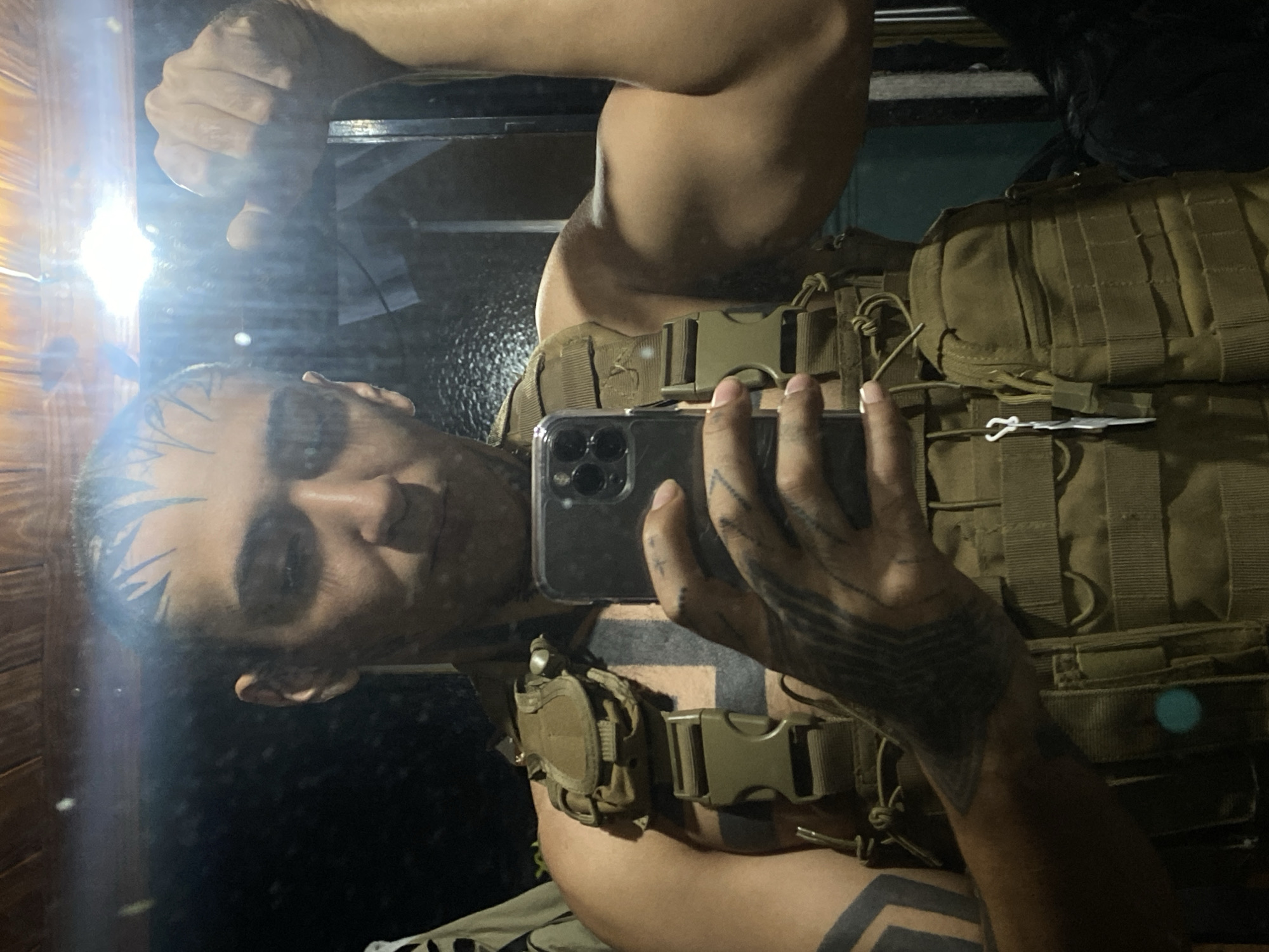 A person takes a selfie in a mirror while wearing a tactical vest with pouches and buckles. The person has tattoos on their head, arms, and chest. The background is blurry and dark, but it appears to be an indoor location. The location is not identifiable as Buenos Aires, Argentina.