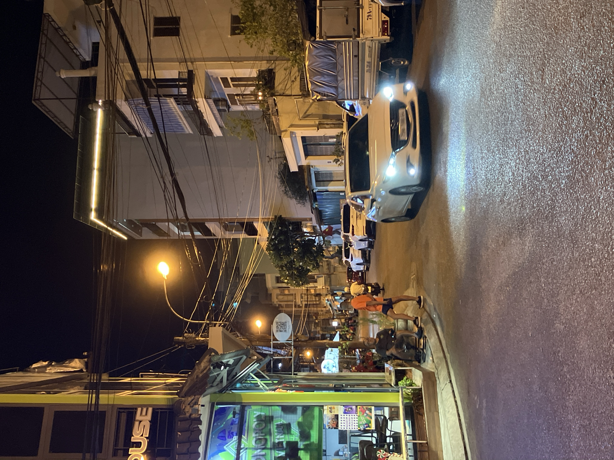 The image depicts a street scene at night in Nha Trang, Vietnam. A white sedan is driving on the right side of the road, its headlights illuminated. A covered truck is parked to the right of the sedan, with visible license plates that appear to be Vietnamese. On the left side of the street, a shop with neon lights is visible, displaying text that includes "MONOPOLY VIETNAM". Two men are seated on a low wall or step in front of the shop. One man is wearing a dark shirt and the other an orange shirt. Further down the street, other vehicles and buildings are visible, illuminated by streetlights and building lights. The overall impression is of a bustling commercial street at night.