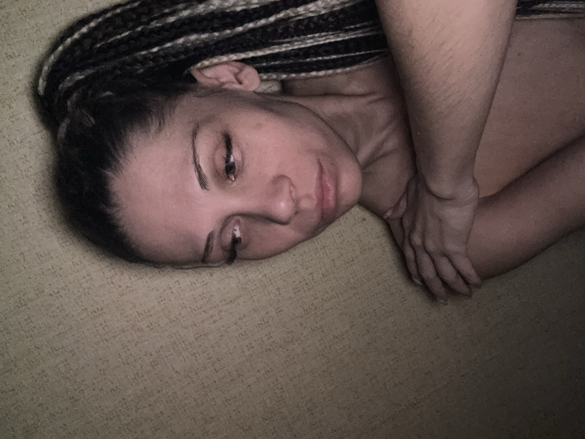 A person with braids is lying on a textured, light-colored surface. The person's head is towards the upper left, and the body extends diagonally across the frame. Their arms are crossed over their body. The lighting is dim. The location is not identifiable as Rusanovo, Russia from the image.