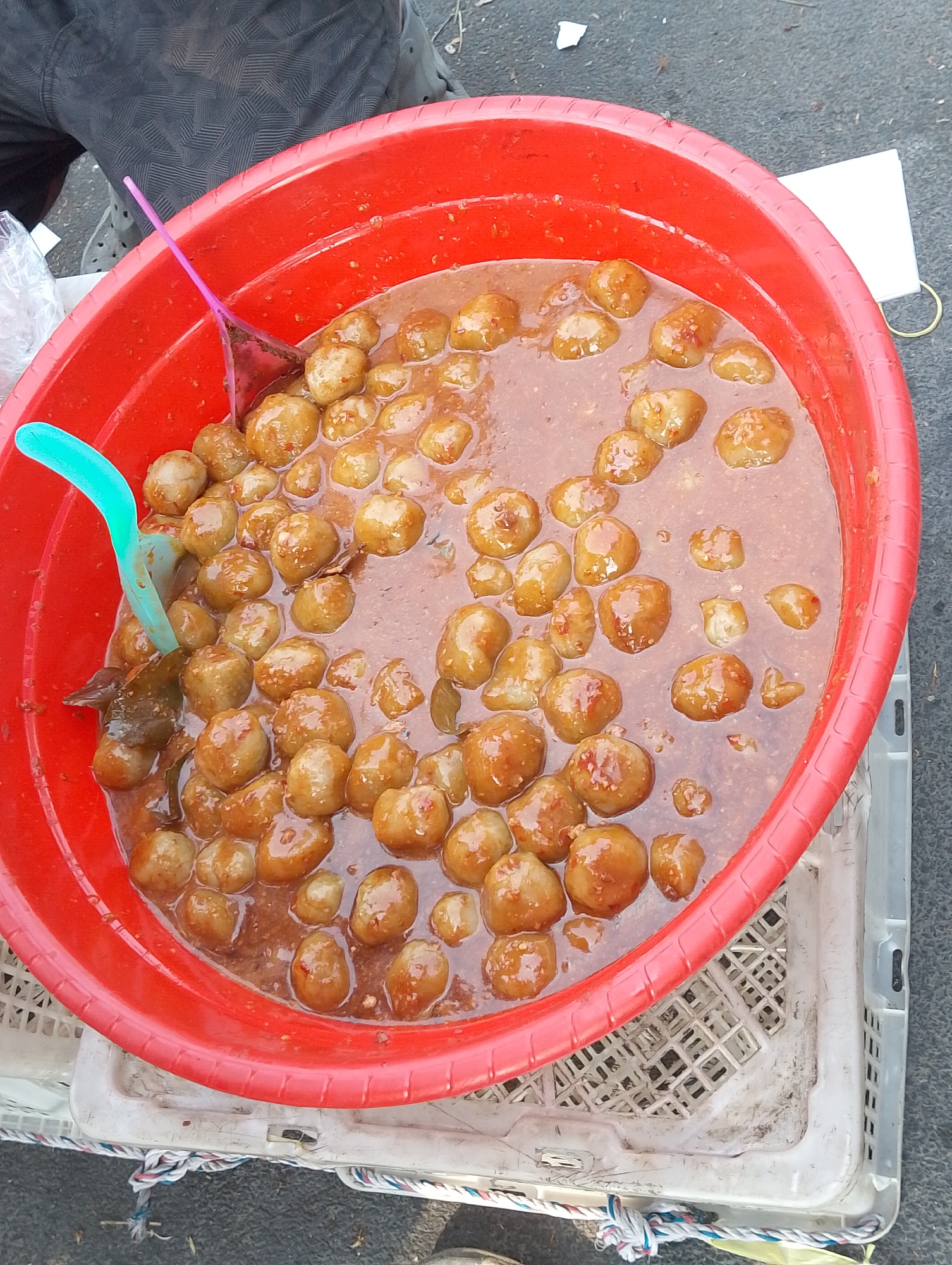 In the city of Karawang, Indonesia, a bright red plastic basin overflows with small, round, brownish meatballs in a savory sauce. Two plastic utensils, one turquoise and the other pink, rest inside the basin. The basin is placed on top of a white plastic crate, which is then situated on a gray, textured surface, possibly a sidewalk or road. The background shows the lower portion of a person's legs, wearing dark patterned trousers. The scene appears to be an outdoor food stall or market setting.