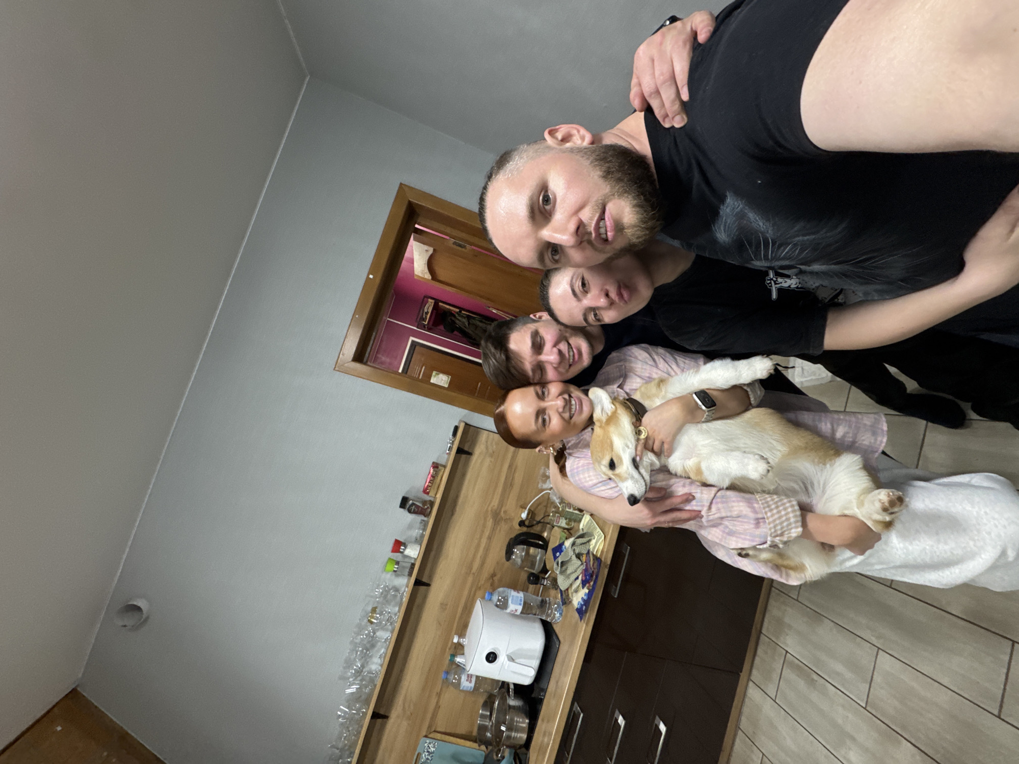 A selfie taken in a kitchen of what appears to be four people and a corgi dog. The people are stacked close together, smiling at the camera. The kitchen has light gray walls and wooden cabinets. A door is partially visible in the background. Objects on the countertop include glassware, a coffee maker, and bottled water. The location is likely Chaikovsky, Russia, based on the photographer's prior images.