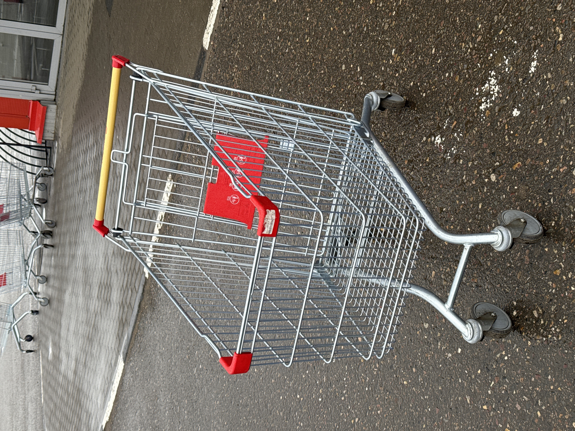 The image shows a shopping cart on wet asphalt, presumably in Ново-Никольское, Russia. The cart is silver metal with a yellow handle and red plastic accents. It sits on the asphalt with other carts behind it, near a building with glass doors.