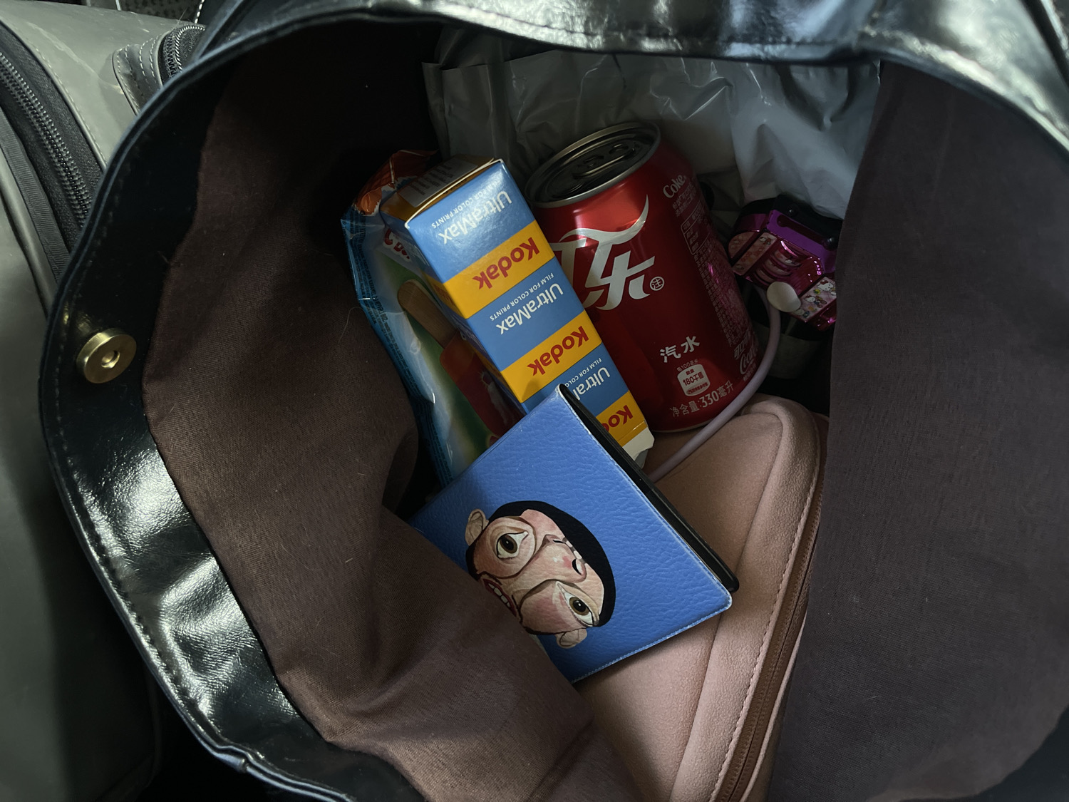 A close-up, overhead shot reveals the contents of a dark bag with a brown fabric lining. The bag holds various items, suggesting casual storage or travel.
Prominently featured is a red Coca-Cola can with its distinct logo. Visible Chinese text on the can includes "汽水" (soda), "净含量:330毫升" (net content: 330 ml), and "180千" (180 kJ or kcal). Stacked next to it are several yellow and blue boxes of Kodak UltraMax film, clearly labeled with "Kodak", "UltraMax", and "FILM FOR COLOR PRINTS".
Other items inside the bag include a small blue object adorned with a cartoonish face featuring large eyes and an exaggerated expression, a soft pink or beige zippered pouch, and a small purple object that appears to be an electronic accessory with a white tip. The bag itself has a dark, possibly leather, exterior with a magnetic snap closure visible on the rim. On the left side of the frame, part of a grey zippered bag or surface is visible, implying the bag might be placed inside another container or in a travel setting.
No specific activity is taking place, and the image offers no cues regarding the time of day, weather, or any discernible emotions.